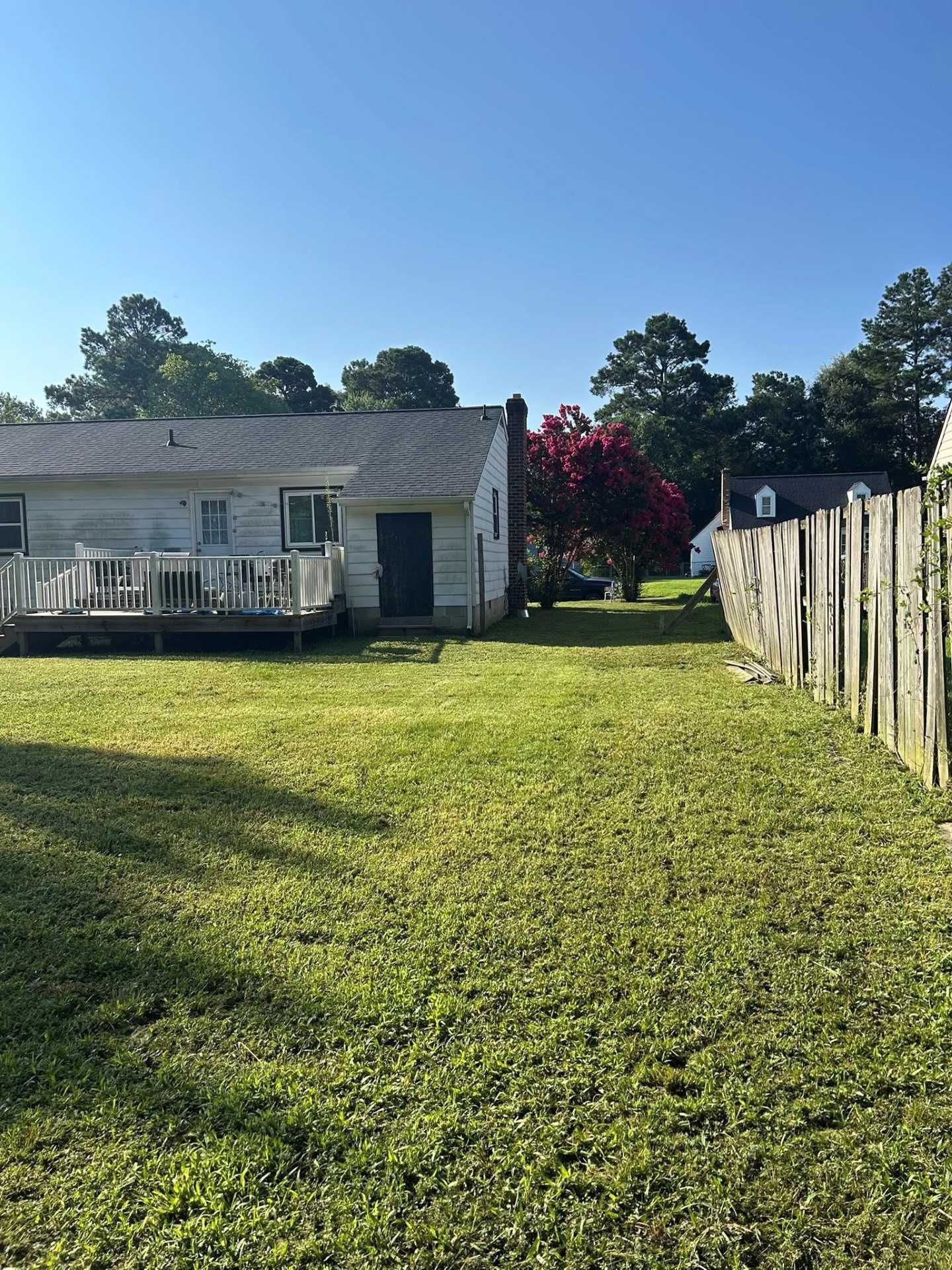 Backyard with house, deck, grass, wooden fence, and trees under a clear blue sky.