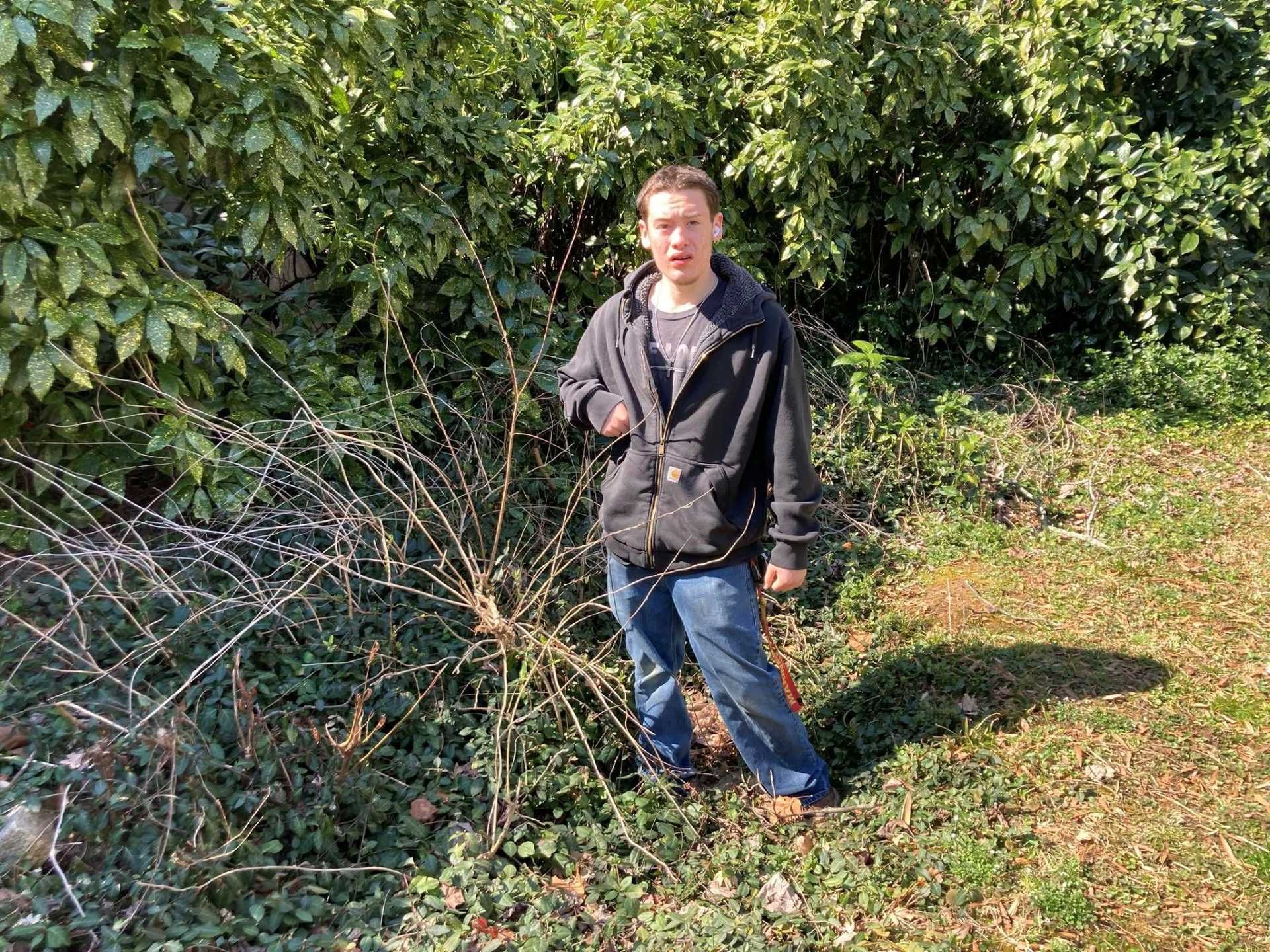 Person in black hoodie and jeans standing outdoors by bushes.