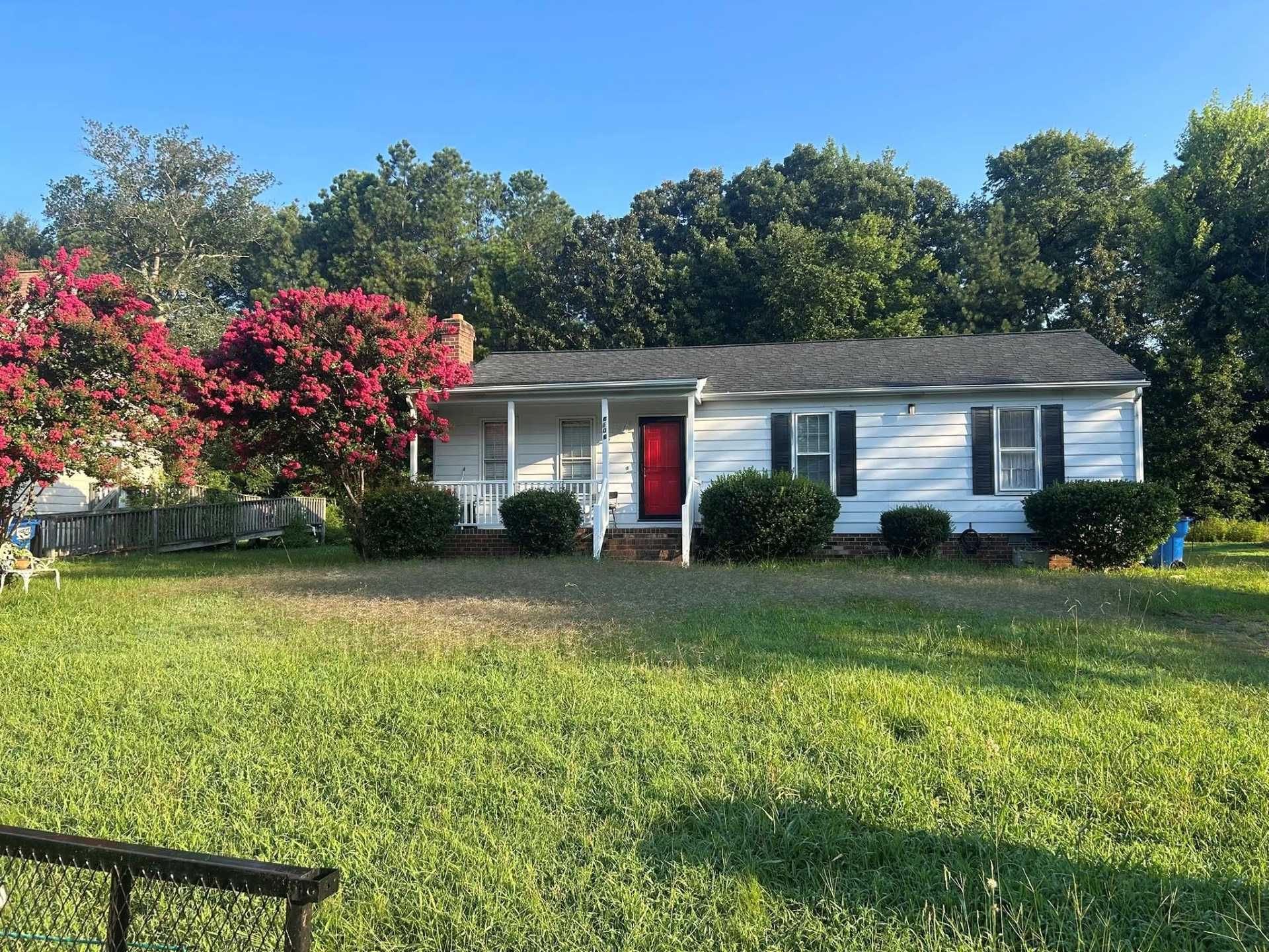 White house with red door, black shutters, and overgrown lawn. Flowering tree to the left.