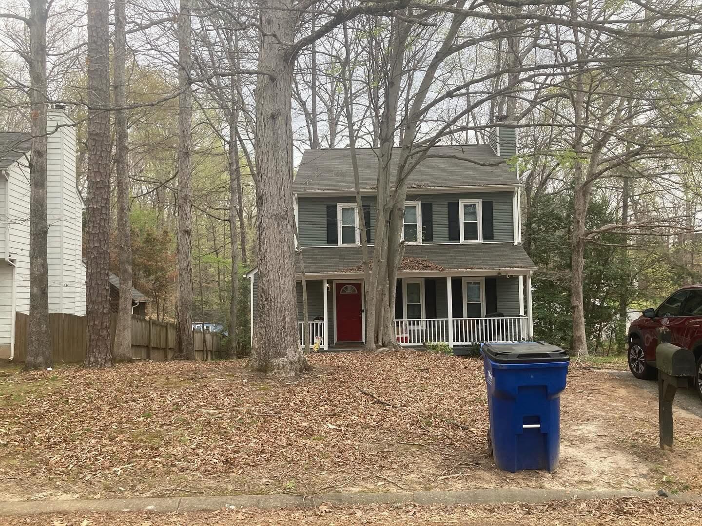 Two-story house with green shutters, red door, and porch. Blue trash bin in front.
