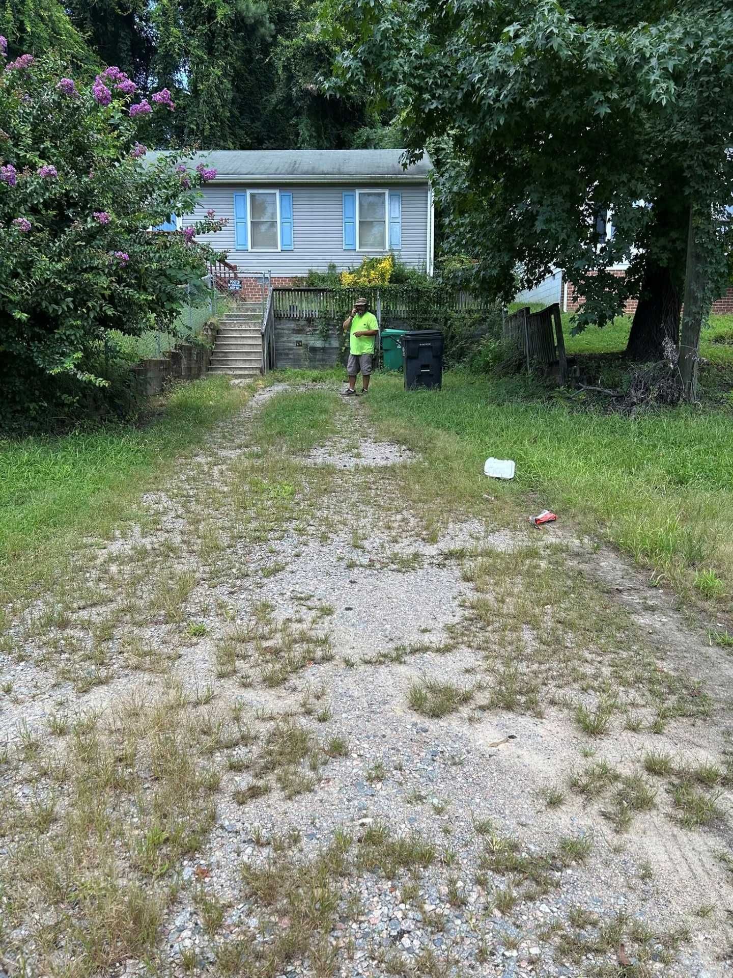 Gravel driveway leads to a small house with blue shutters, a person stands nearby holding a trash can.