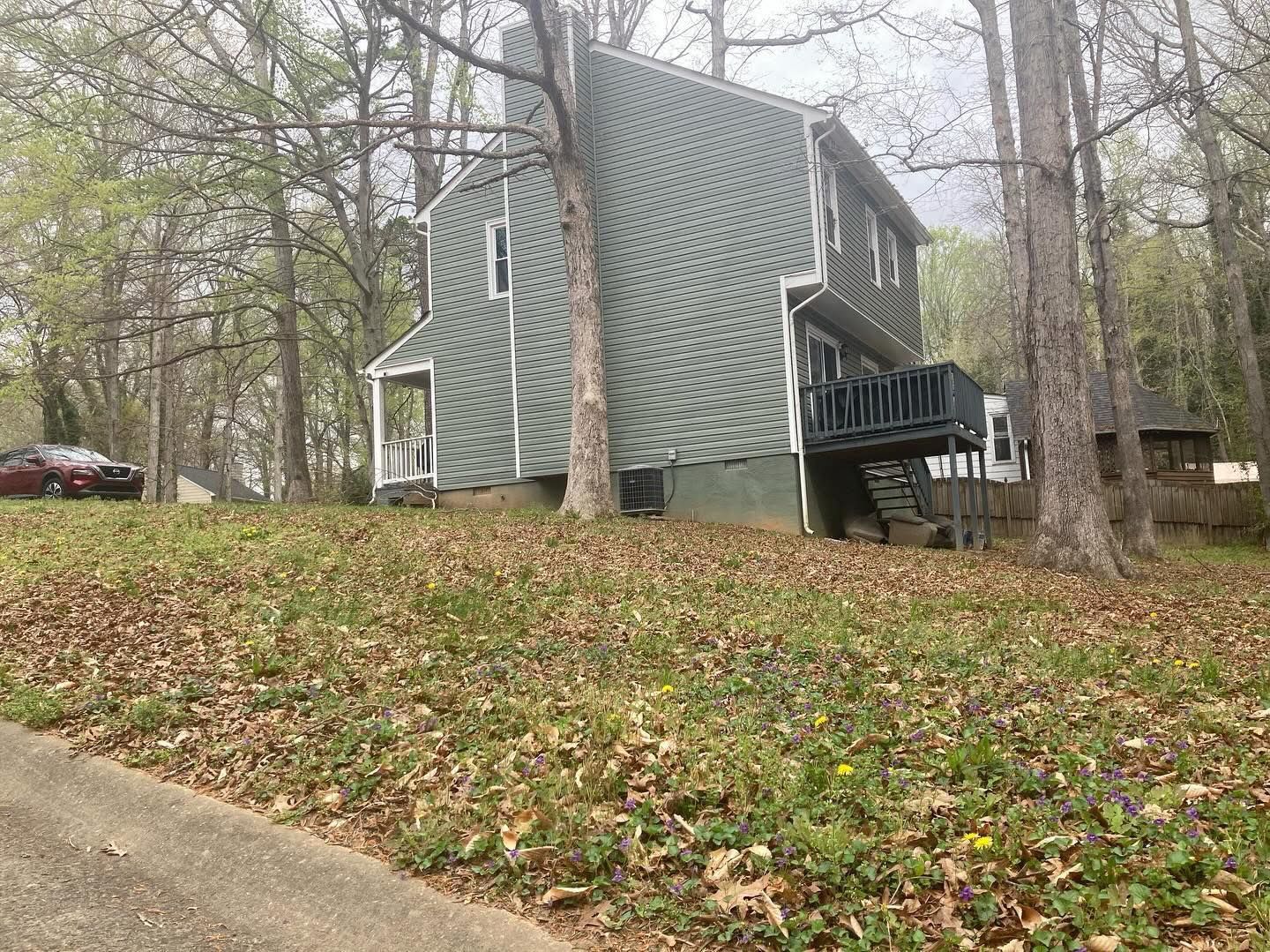Green A-frame house on a grassy hill, surrounded by trees. A car is parked on the left.