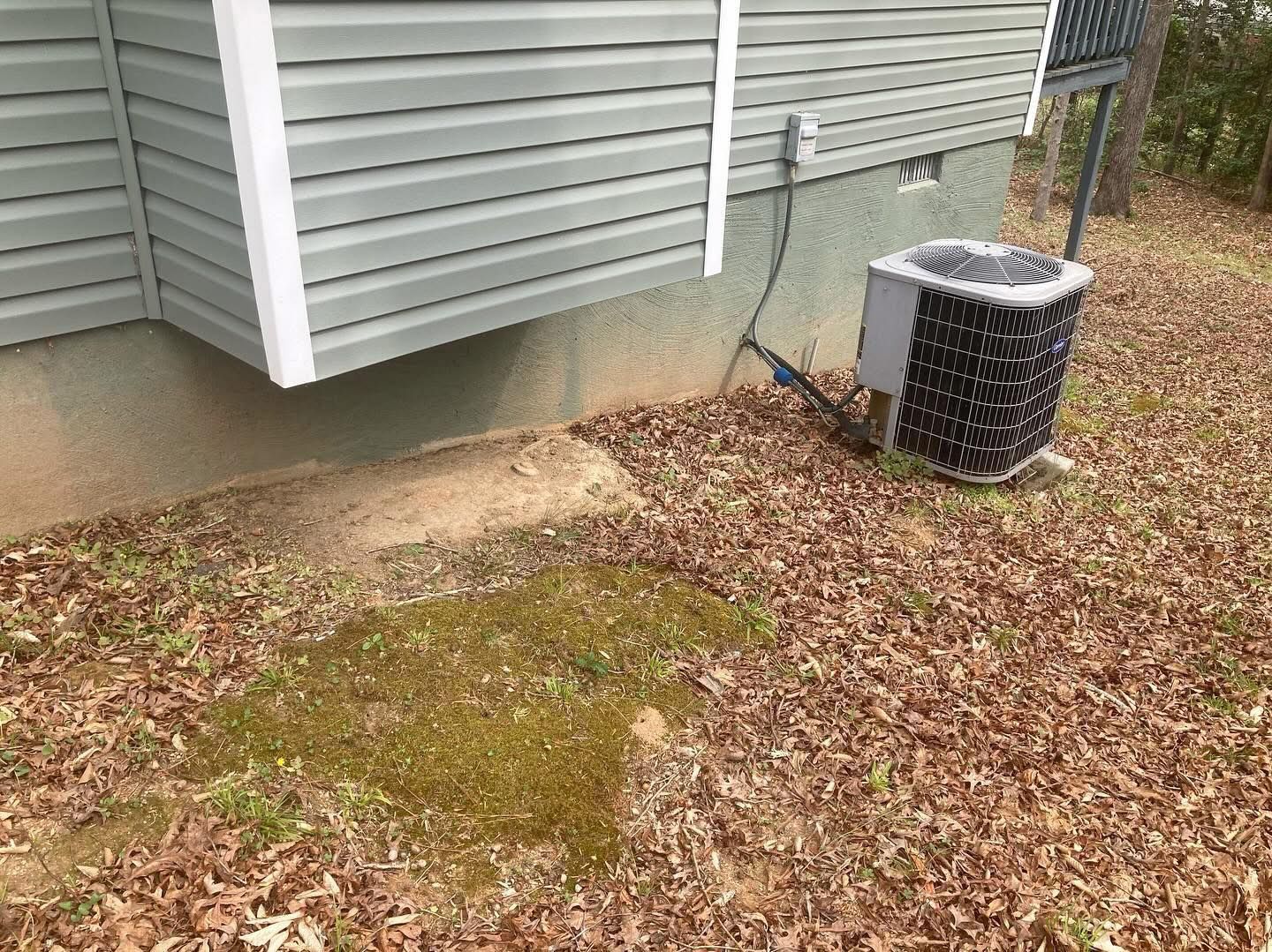 Air conditioner unit beside a house; brown leaves and mossy ground.