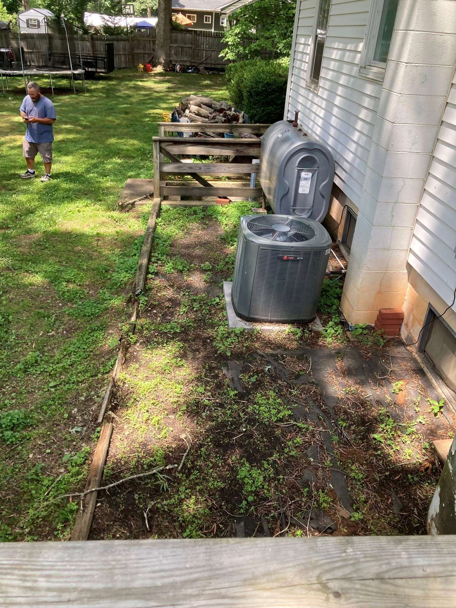 Backyard with air conditioner unit and weeds. A person stands in the grass. Wooden deck in foreground.