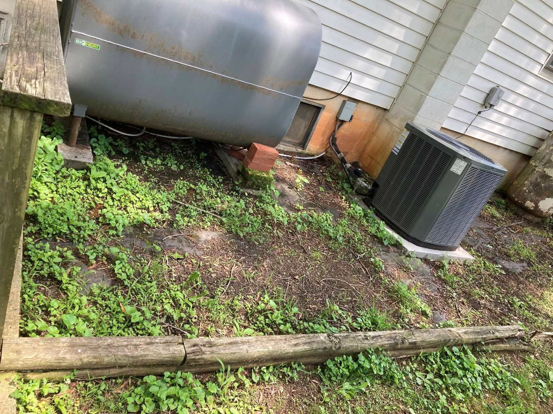 Oil tank and AC unit on a sloped, weedy, wooden-edged lot next to a house.