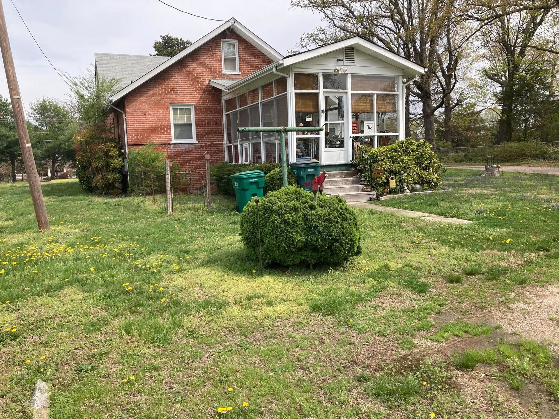 A one-story brick house with a screened porch and green trash cans sits on a grassy lot.