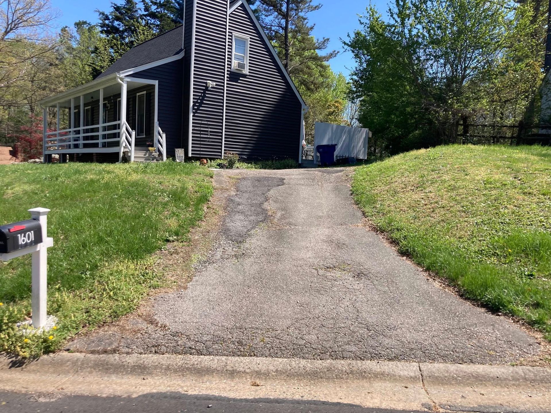 Driveway leading to a dark A-frame house; green grass surrounds the asphalt path, sunny day.