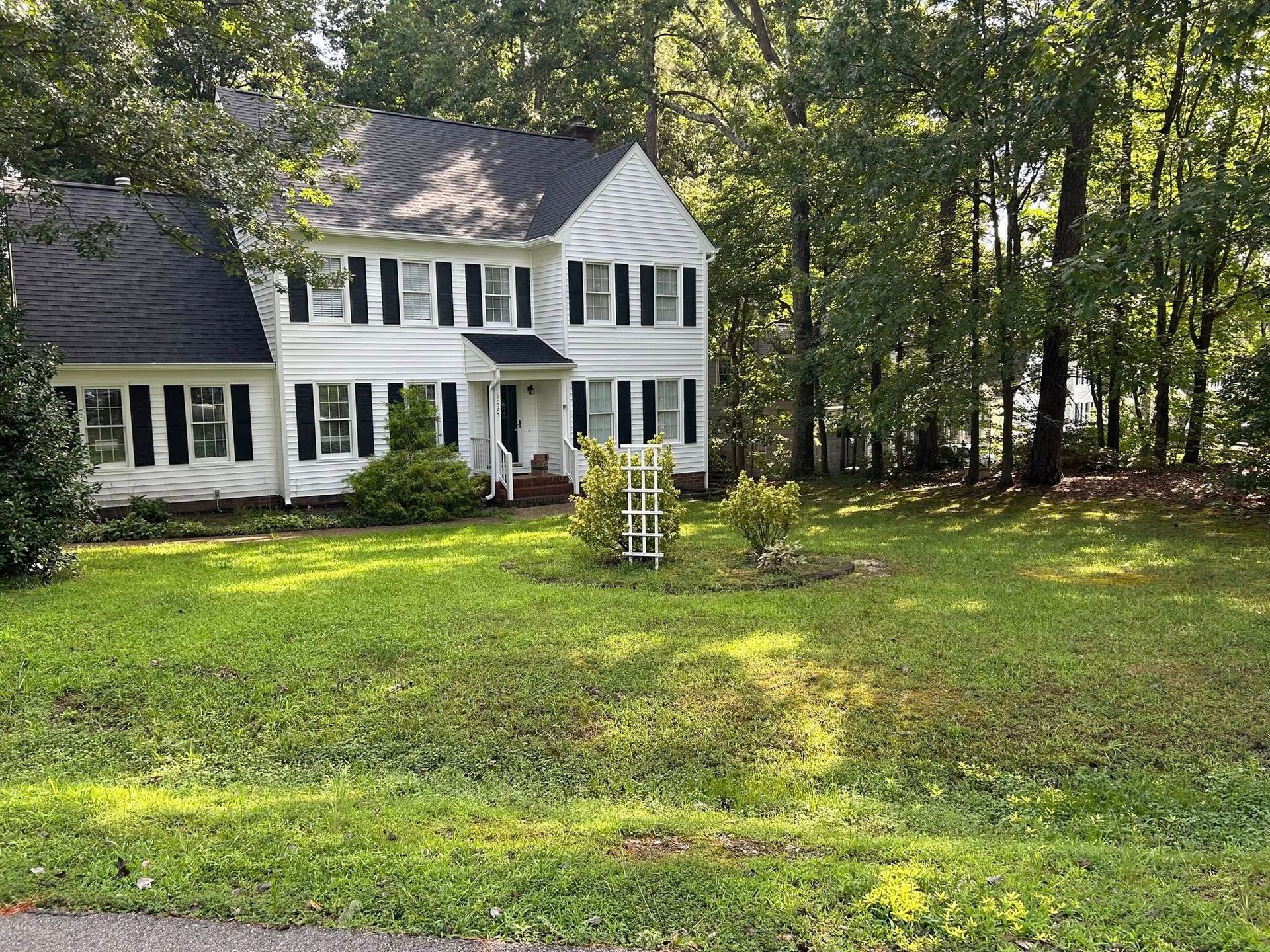 White two-story house with black shutters, on a grassy lot surrounded by trees.