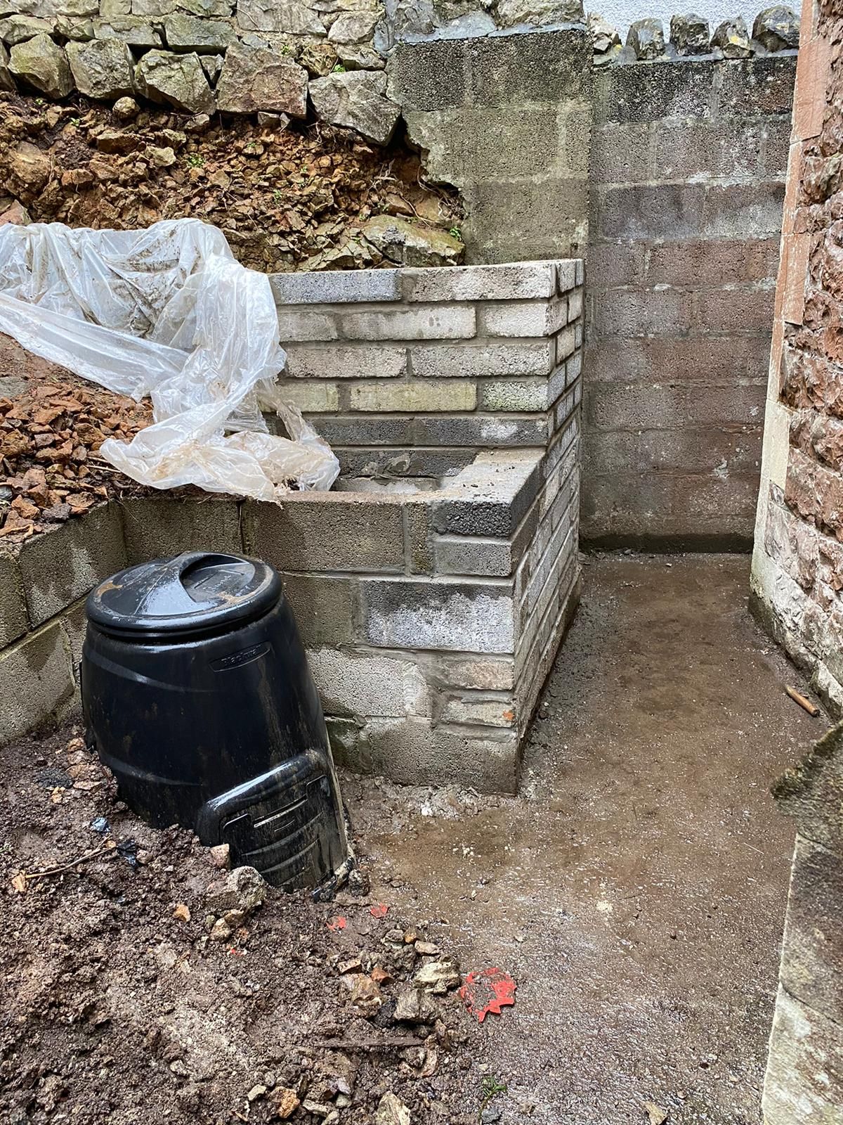 A black barrel is sitting in the dirt next to a brick wall.
