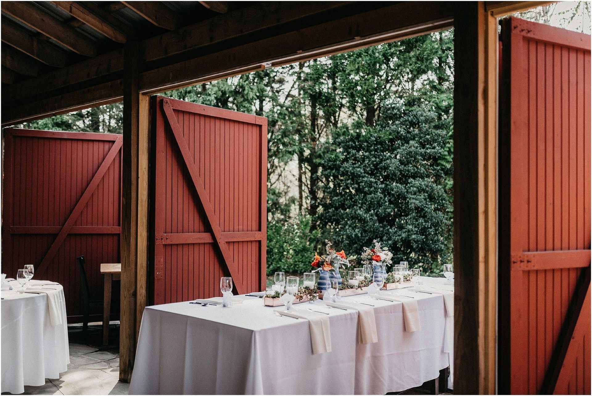 A long table with a white table cloth is under a covered area with red doors.