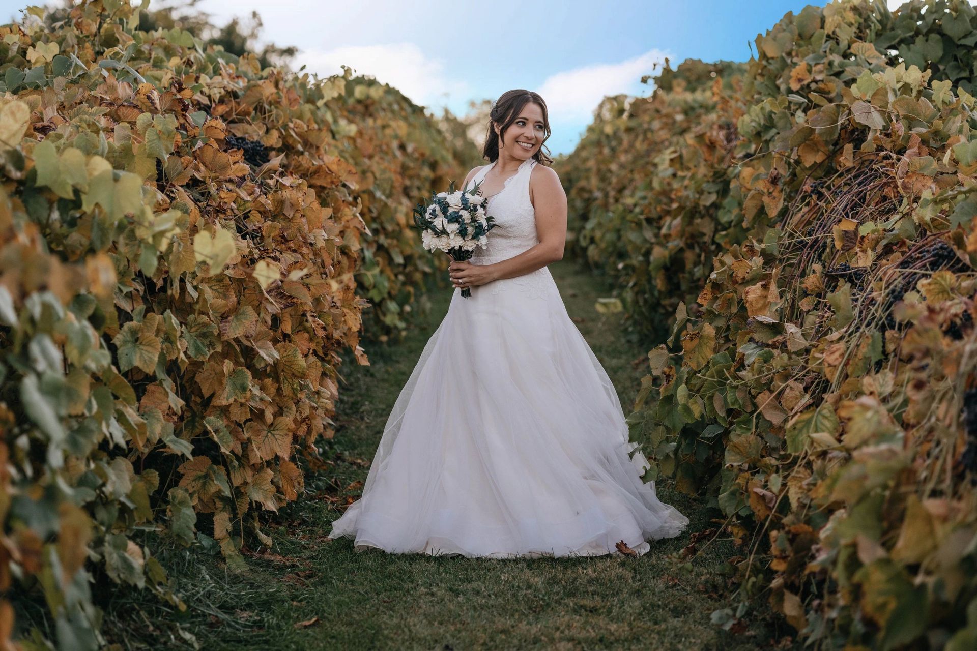 A bride in a wedding dress is standing in a vineyard holding a bouquet of flowers.