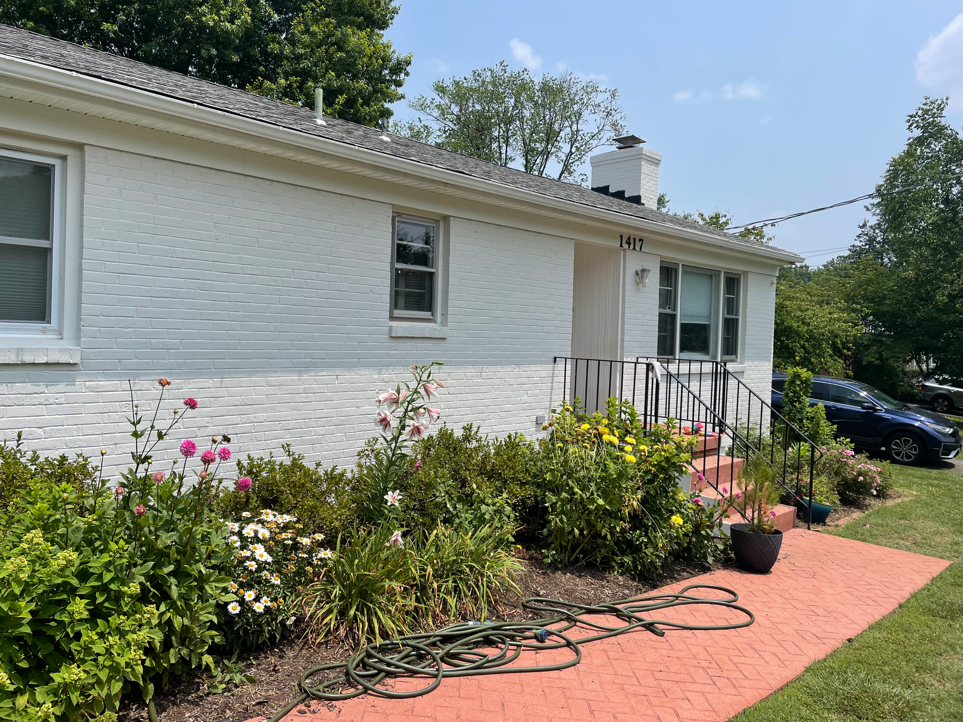 White brick house with a flower garden and red brick walkway. A car is parked on the right.