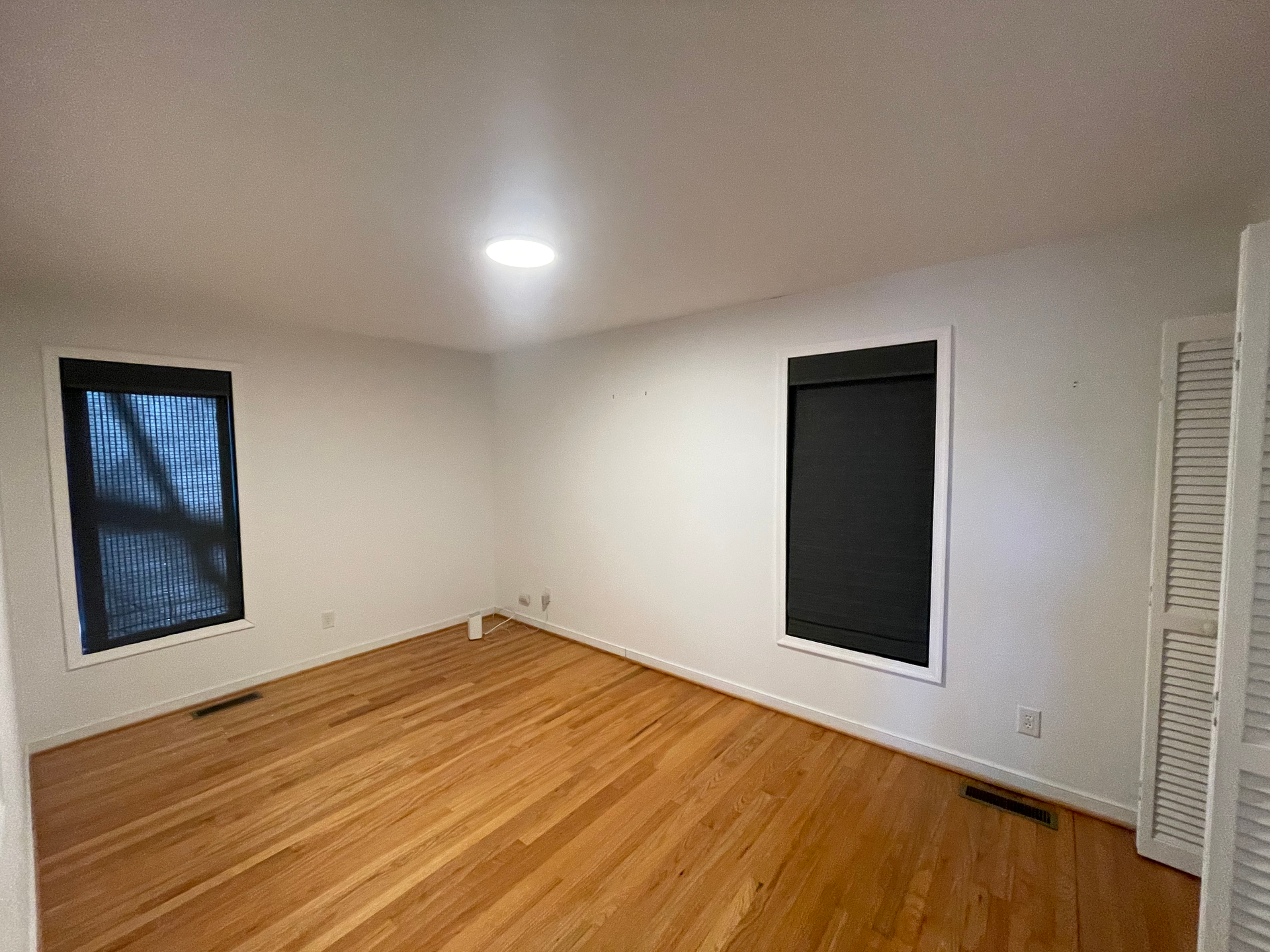 Empty room with hardwood floors, white walls, two dark window shades, and a skylight.