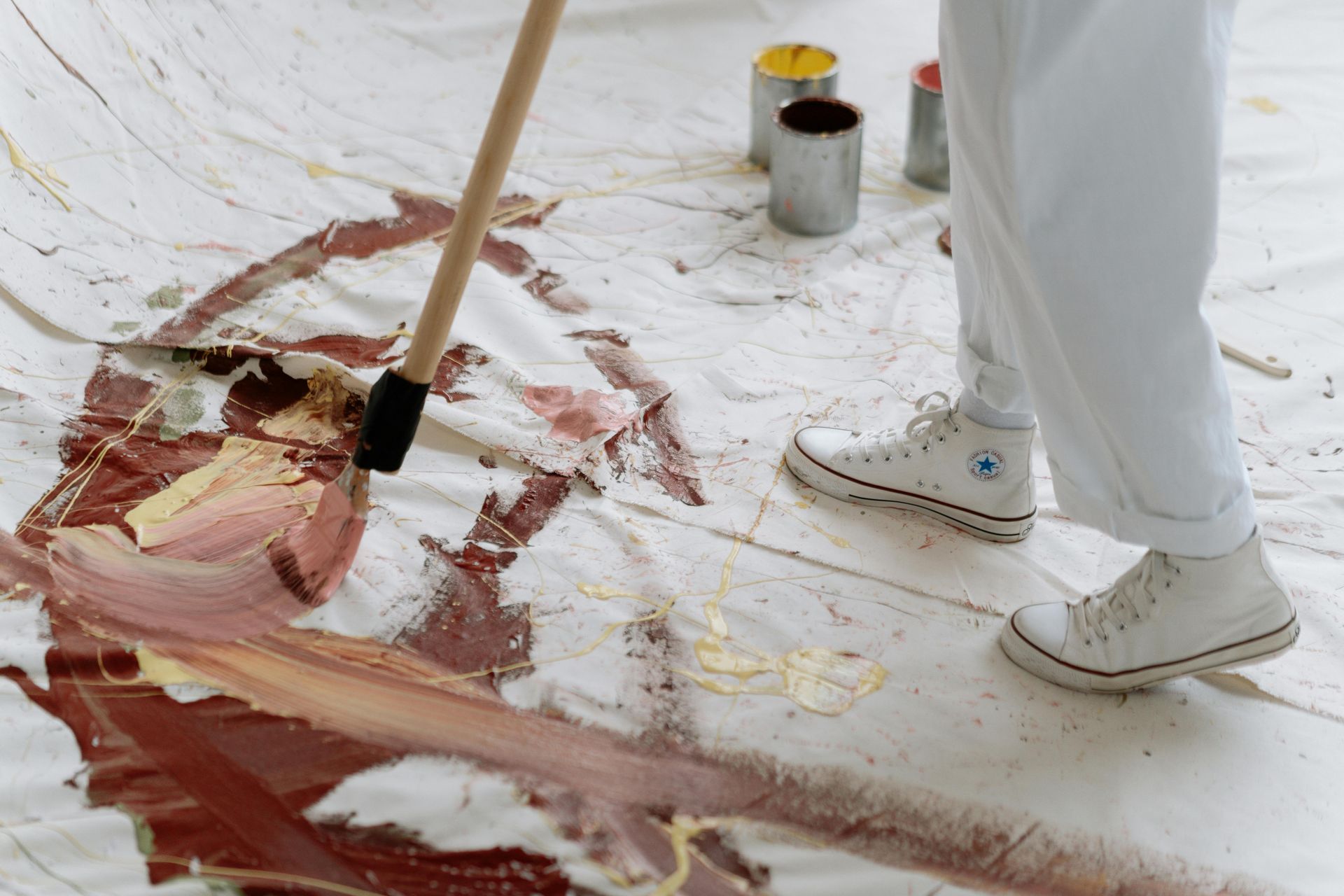 Artist painting on a large canvas, wearing white pants and sneakers. Paint cans in background.