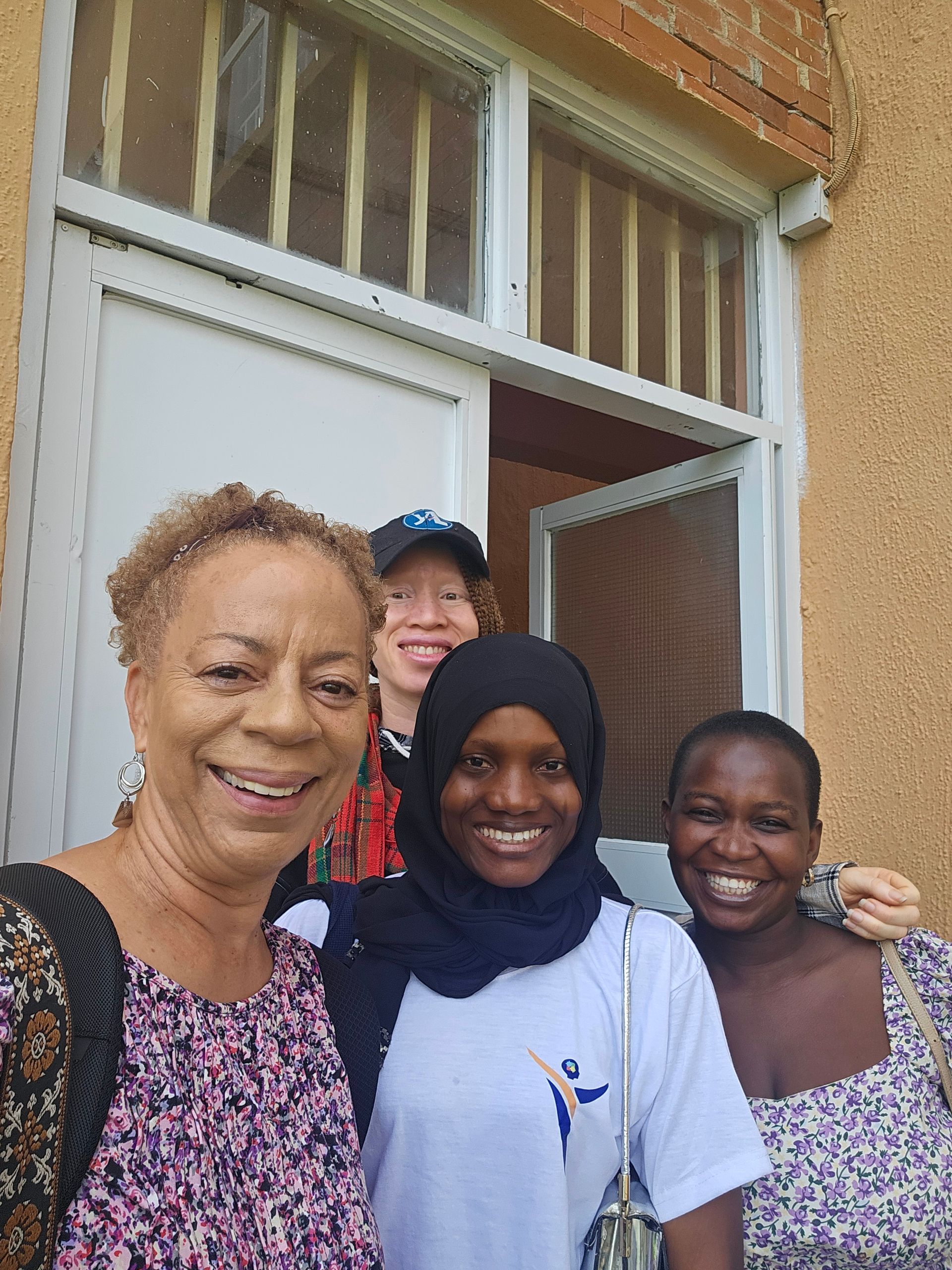 Four people smiling near a door. One woman has a backpack, another wears a hijab, another a blue hat.