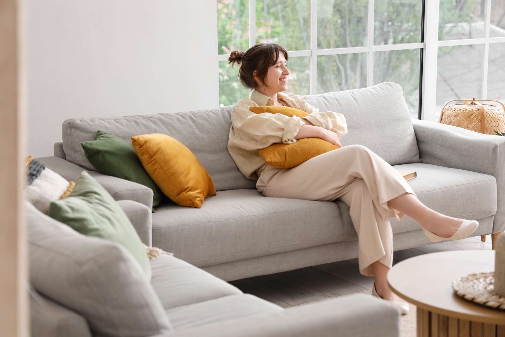 Woman relaxing on a grey sofa, holding a yellow pillow, near a window; sunlight streams in.