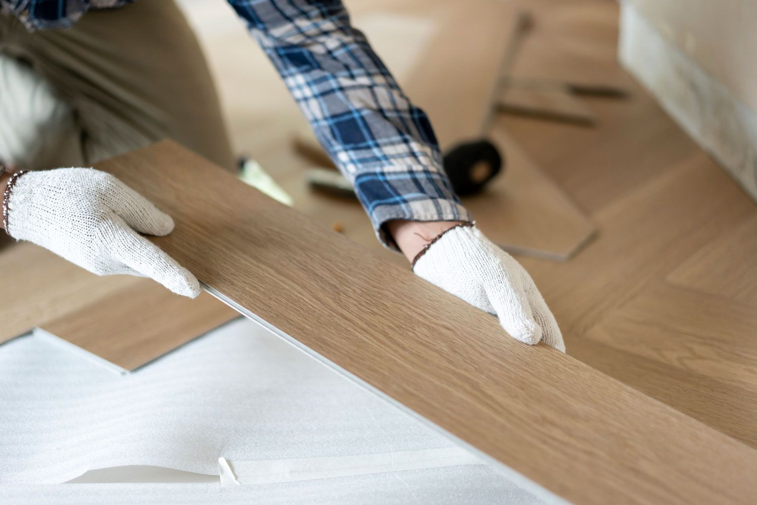 Person installing wood flooring, wearing gloves and plaid shirt.