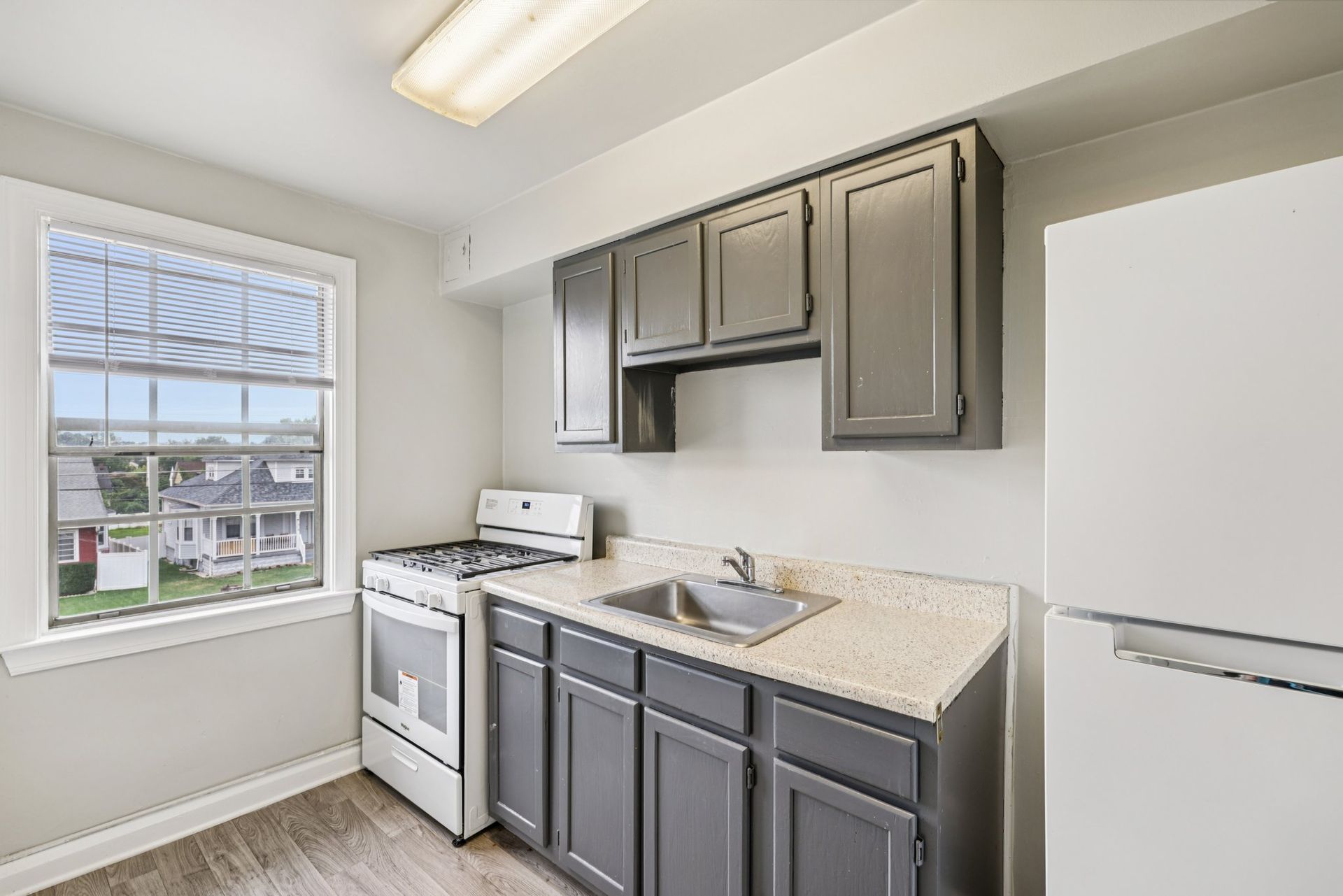 Small kitchen with gray cabinets, white appliances, and a window overlooking a neighborhood.