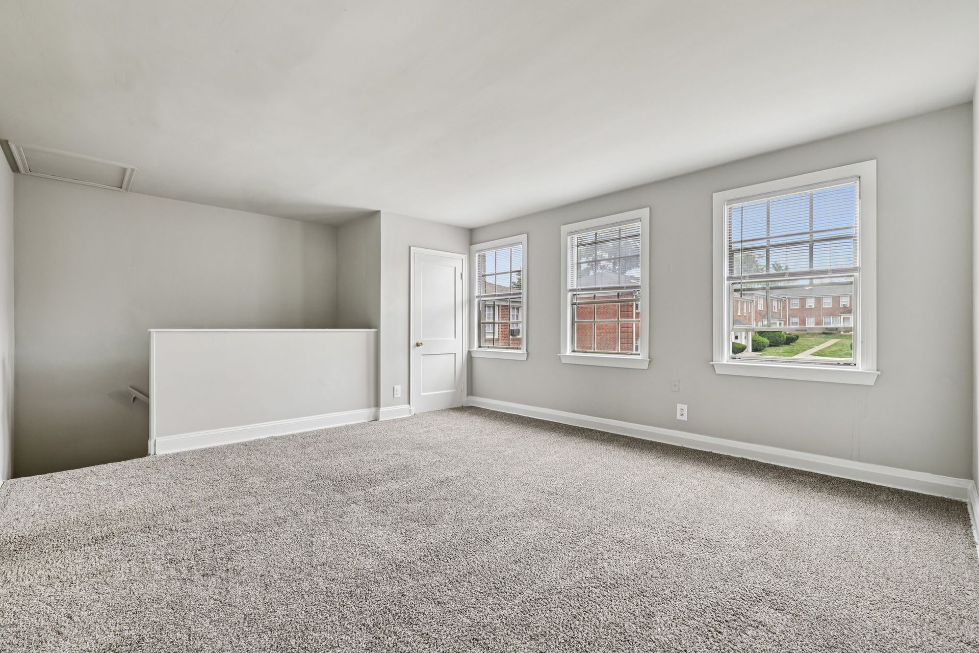 Empty, carpeted room with three windows, white door, and half wall on the left.