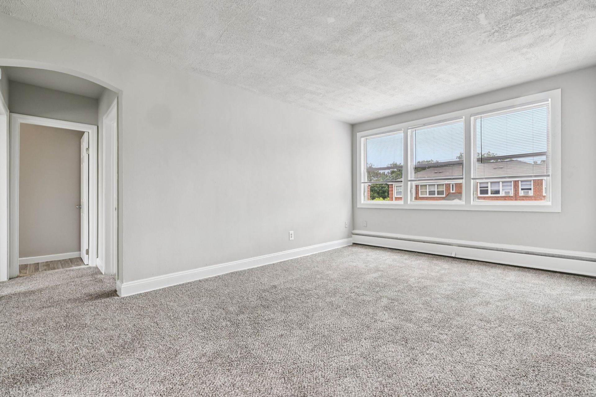 Empty, bright room with gray walls, carpet, and a large window overlooking a residential area.