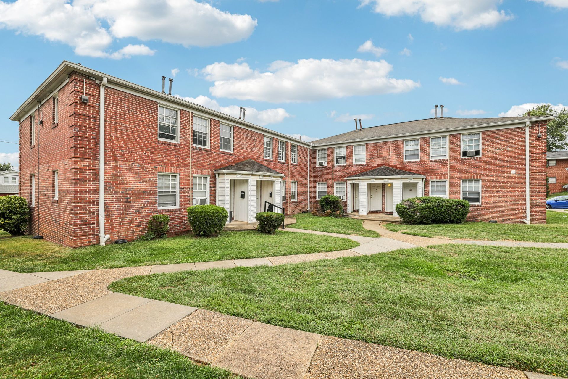 Two-story brick apartment buildings with white doors and windows; green lawn, blue sky, and a brick walkway.