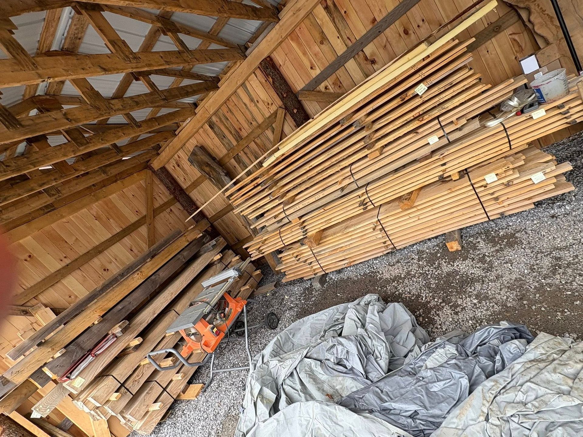 Lumber stacked inside a wooden building with a rafter roof; gray tarps cover the ground.