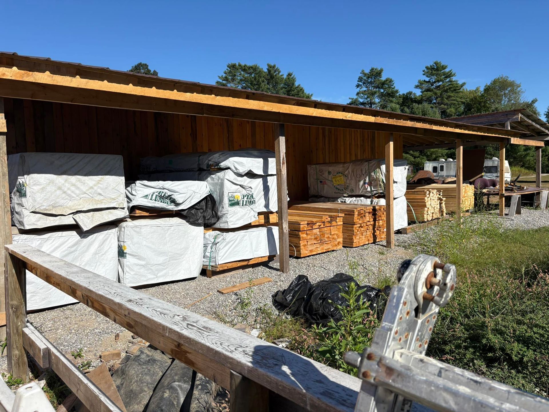 Lumber storage shed with stacks of wood, fence in the foreground, sunny day.