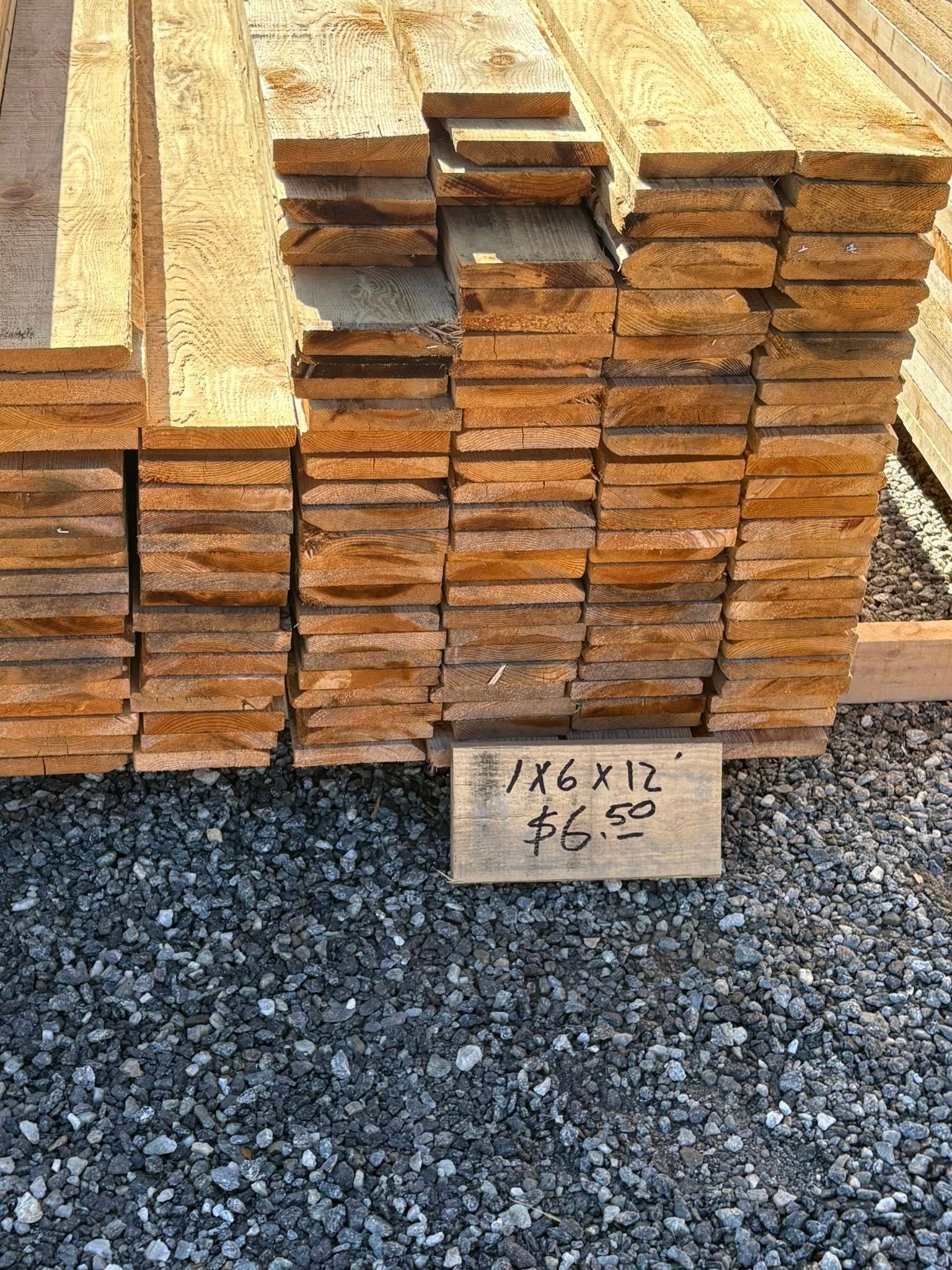 Stack of wooden boards with a price sign that reads on a gravel surface.