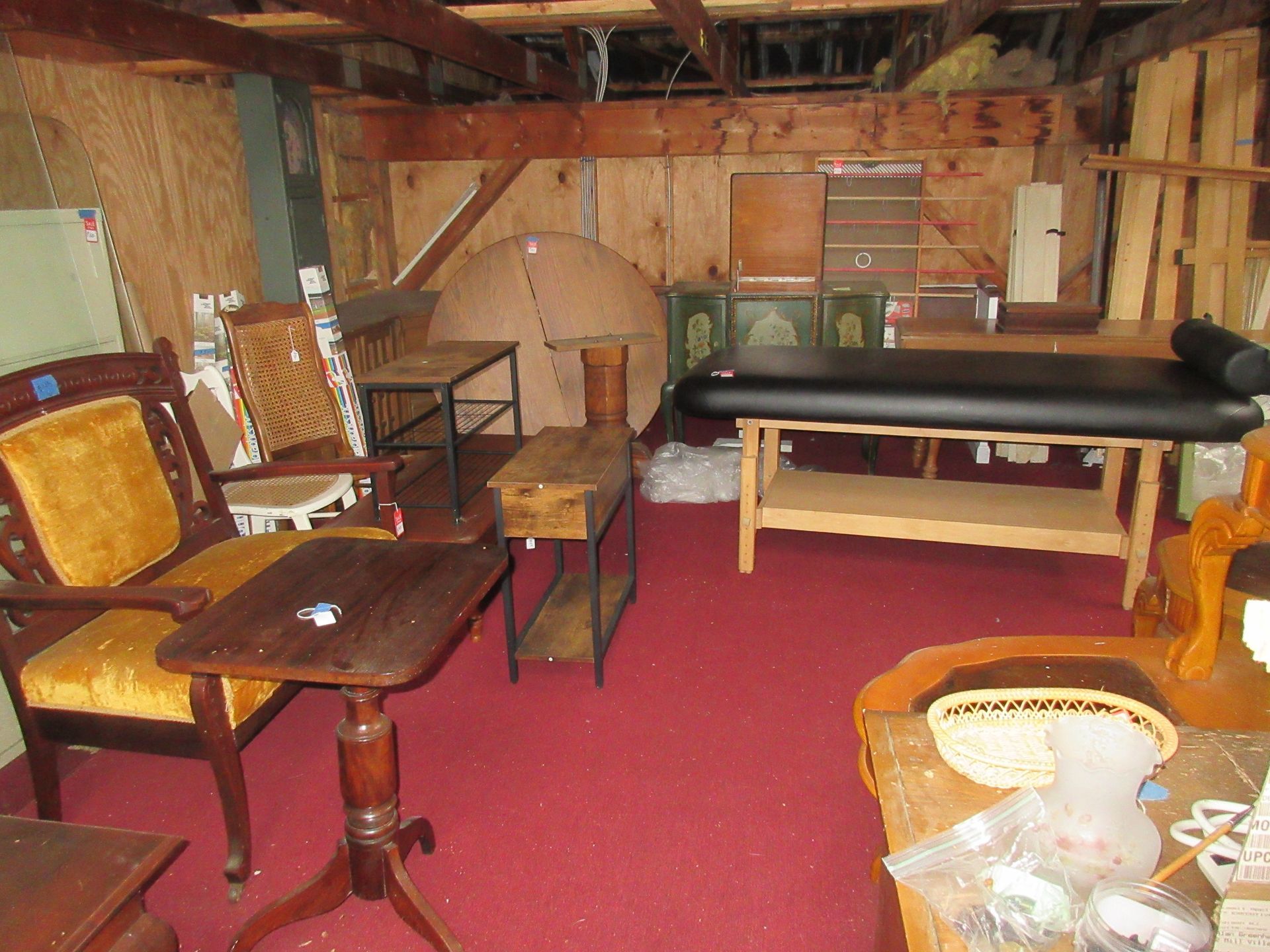Room with various used furniture on a red carpet, including a massage table, chairs, and tables.