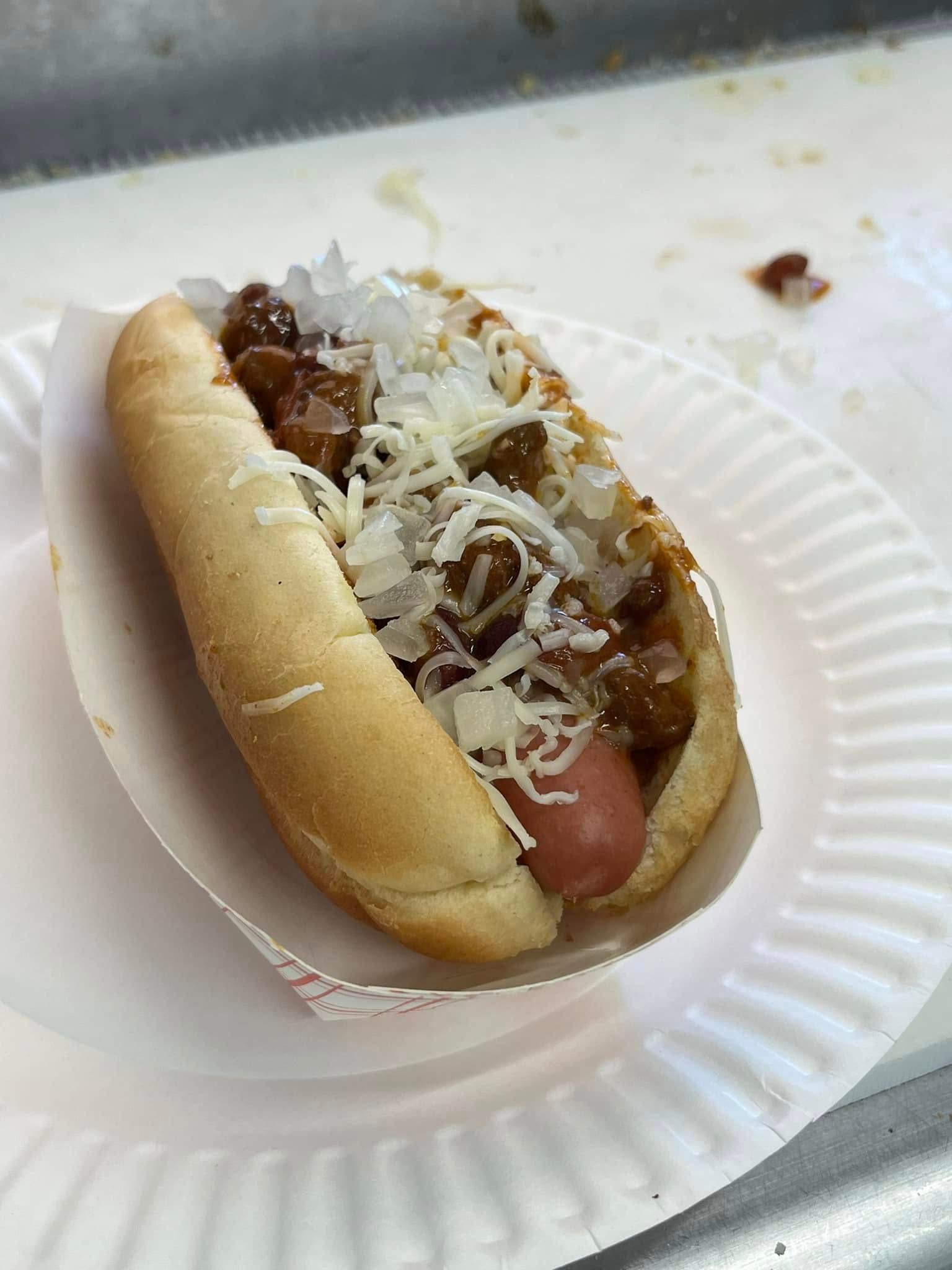 Chili dog in a paper boat on a white paper plate, topped with onion and cheese.