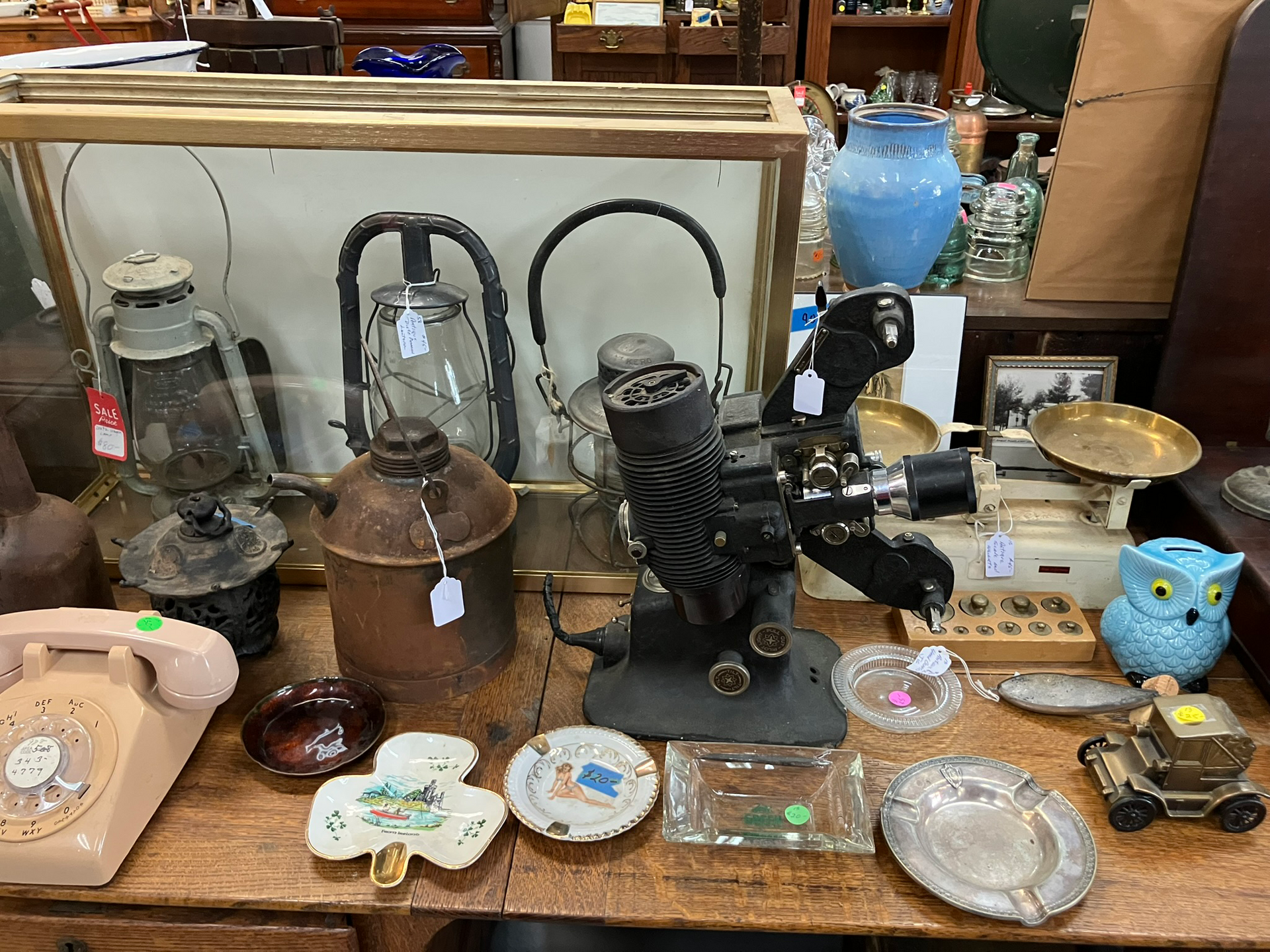 Antique items on a wooden table: lanterns, projector, telephone, owl figurine, dishes, scale.