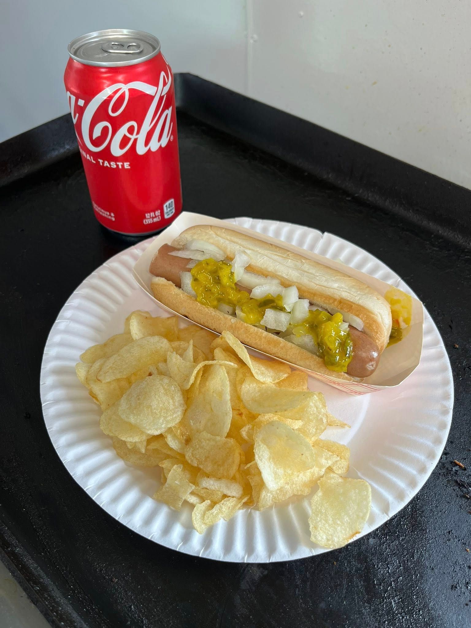 Hot dog with relish and onions, potato chips, and Coke on a paper plate.