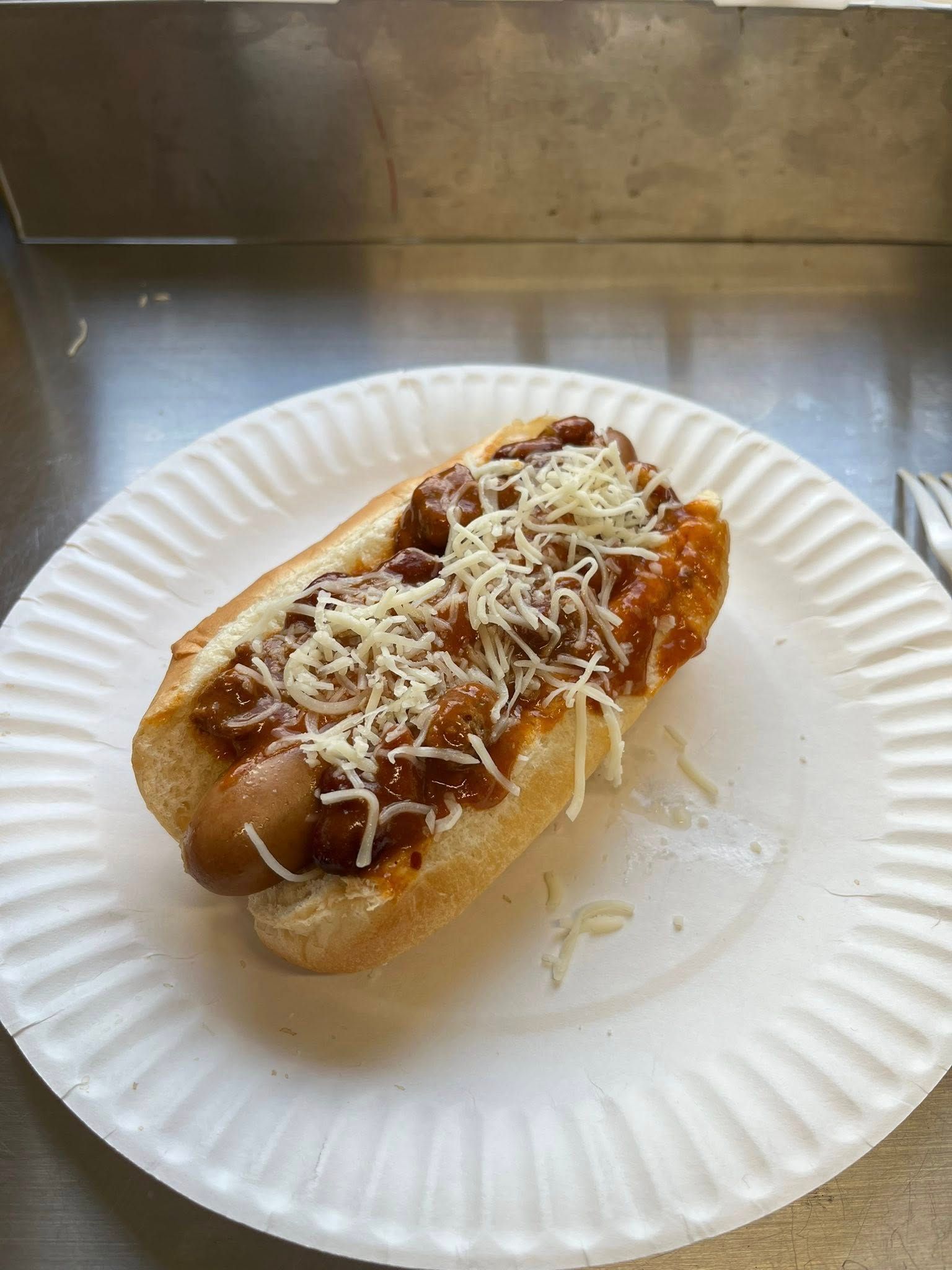 Hot dog on a white paper plate, topped with chili and shredded cheese.