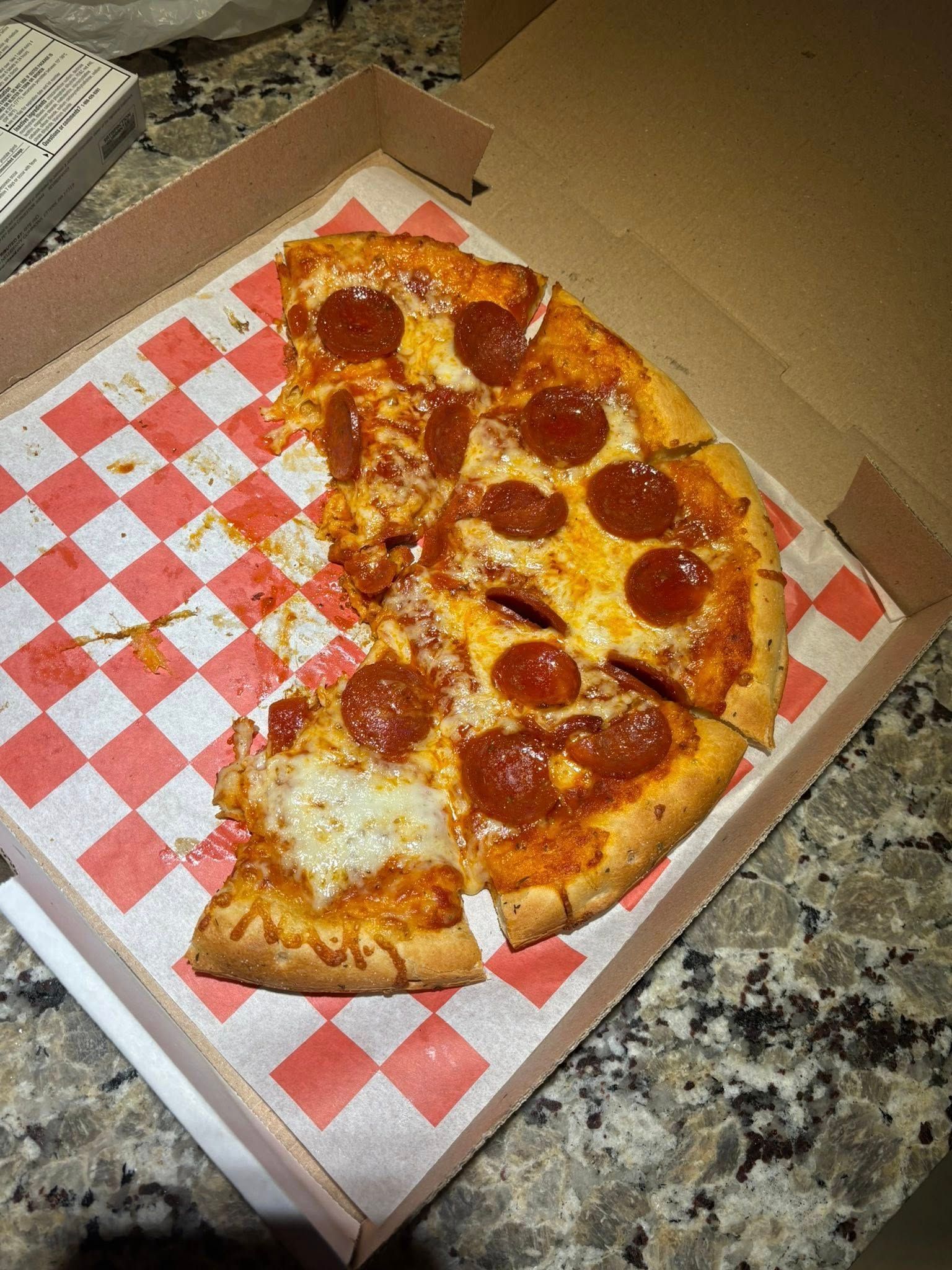 Pepperoni pizza in a cardboard box, partially eaten, on checkered paper.
