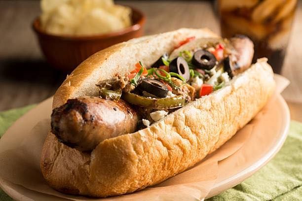 Sausage sandwich in a bun, topped with olives, peppers, and cheese, with chips and a drink.