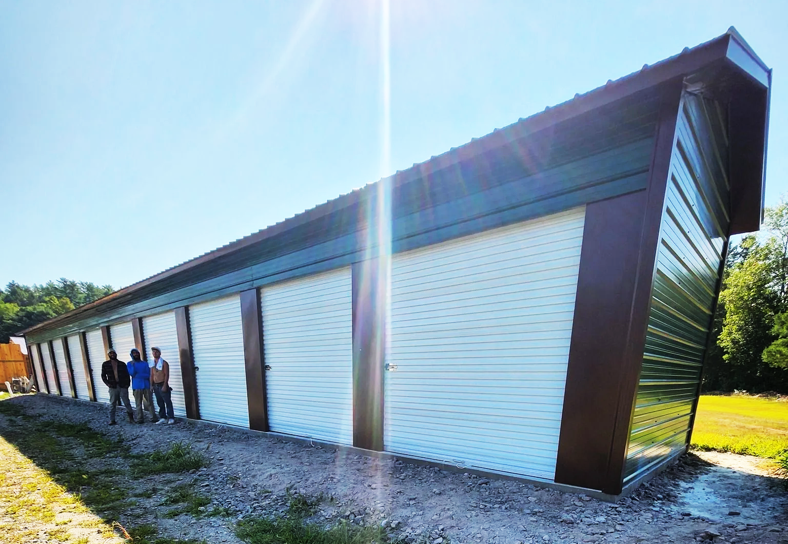 Storage units with white doors, brown trim, and a green roof under a blue sky, with three people standing in front.