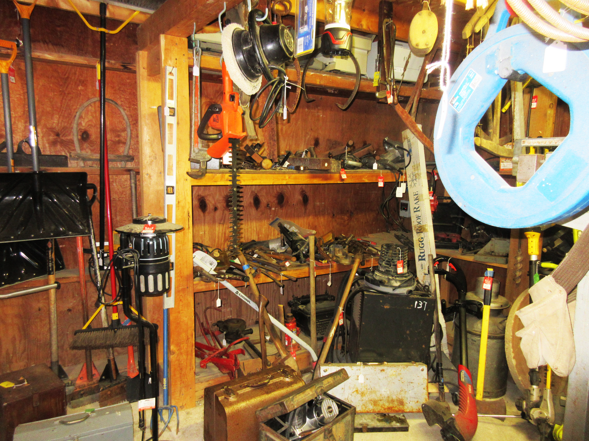Messy tools shed with tools hanging on walls and shelves, various tools on ground.