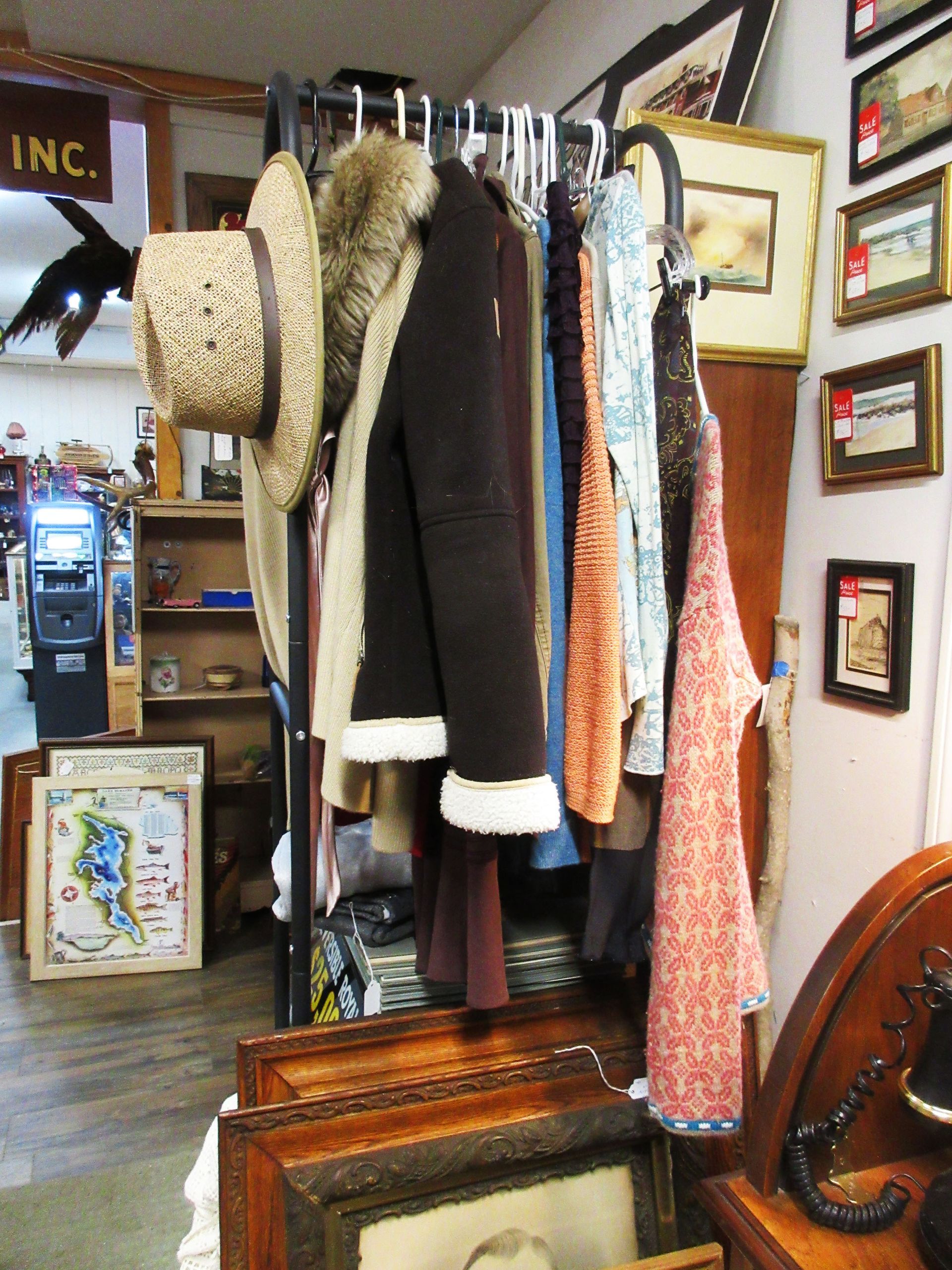 Clothes rack in a thrift store, displaying various jackets and tops; paintings and furniture in the background.