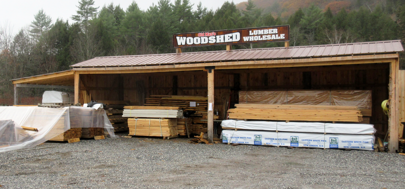 Woods & Co Lumber, lumberyard with stacks of wood under a shed roof, trees in the background.