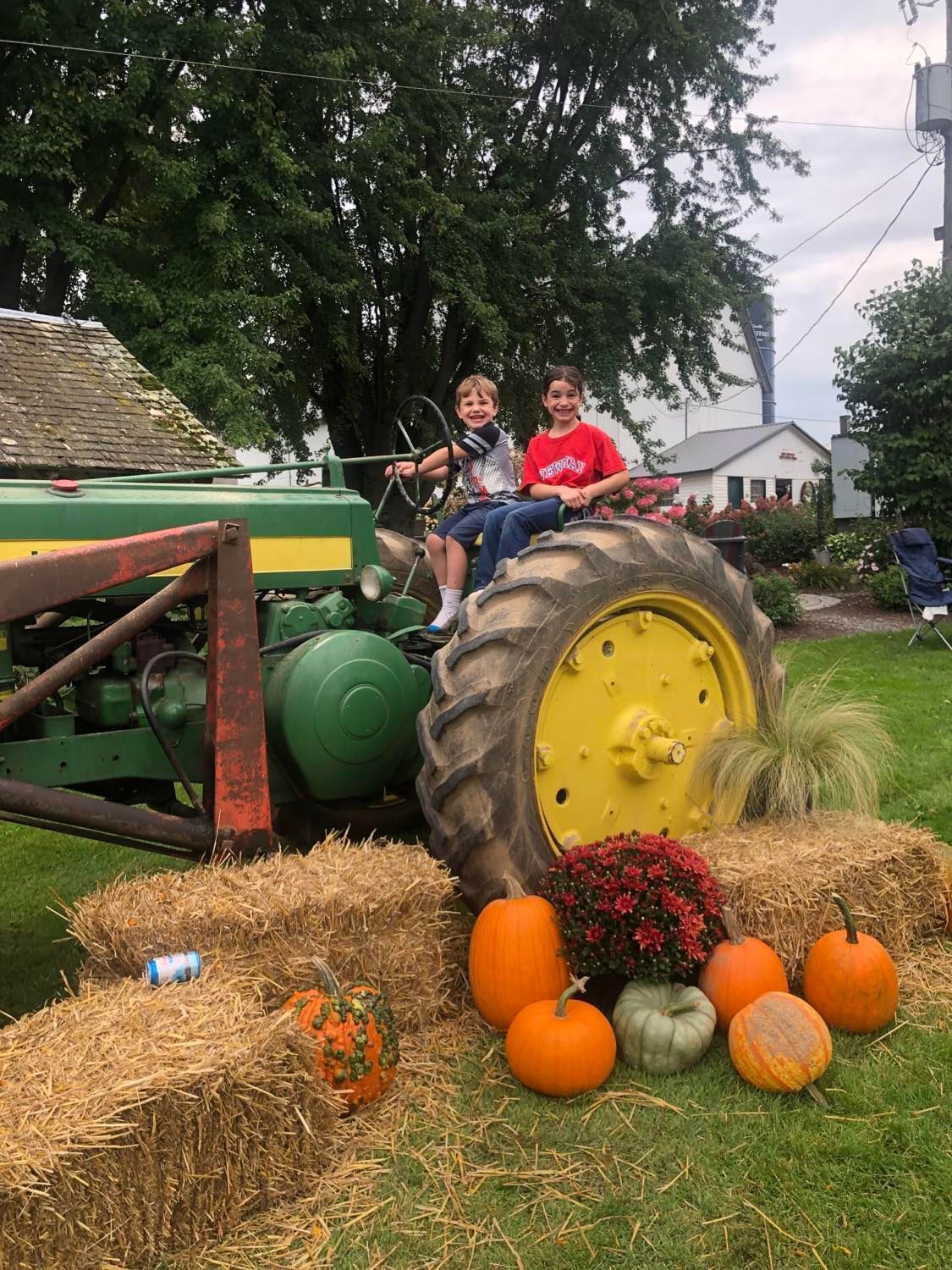 Two children are sitting on a john deere tractor surrounded by pumpkins and hay bales.