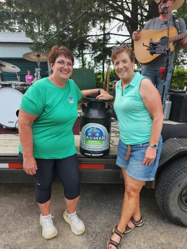 Two women in green shirts stand next to a milk can