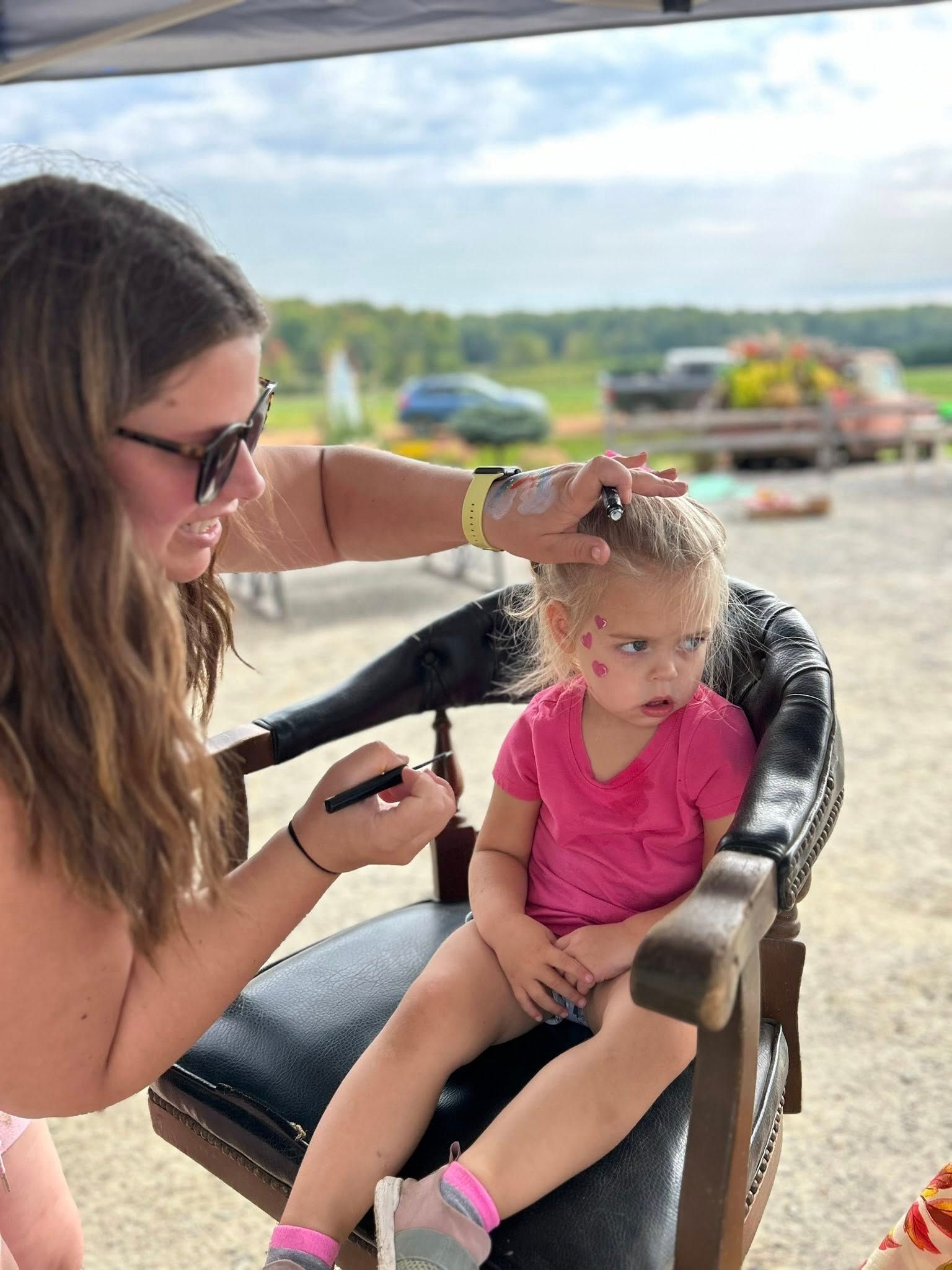 Person applying face paint to a child sitting in a chair outdoors. Child has pink shirt, serious expression.