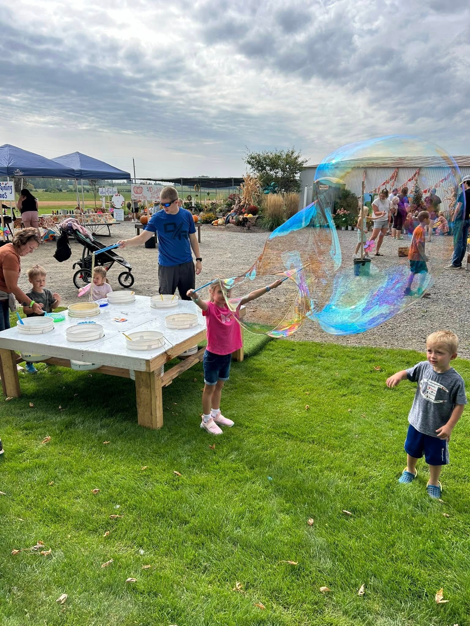 Children playing with giant bubbles at an outdoor event; green grass, tables, tents, and people in the background.