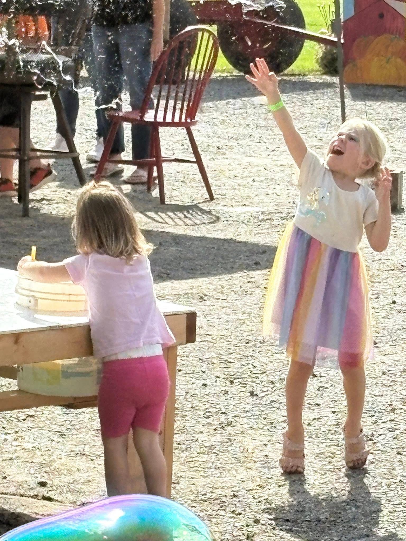 Two young children playing outside; one reaches for bubbles, the other works at a table.
