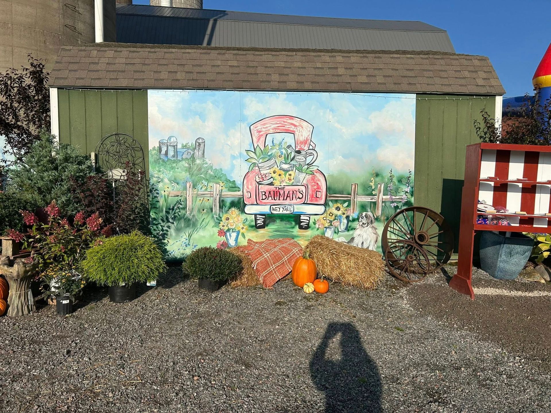 Colorful mural of a red truck carrying flowers, fall décor with pumpkins, hay, and plants, in front of a green shed.