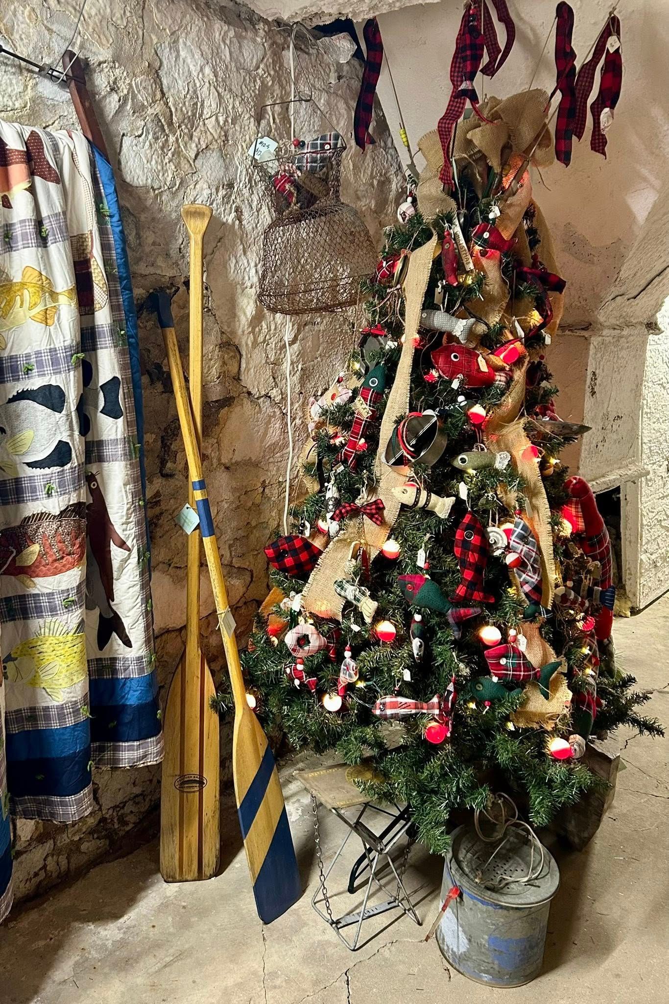 Christmas tree with red ornaments and burlap ribbon, next to wooden oars in a rustic setting.