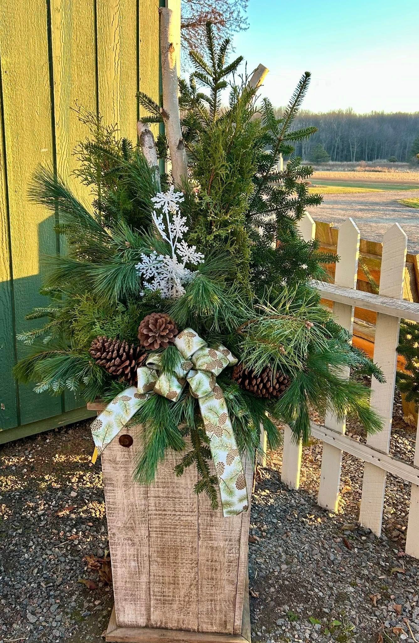 Holiday planter with pine boughs, pinecones, bow, and faux snowflakes. Stands near a white picket fence.