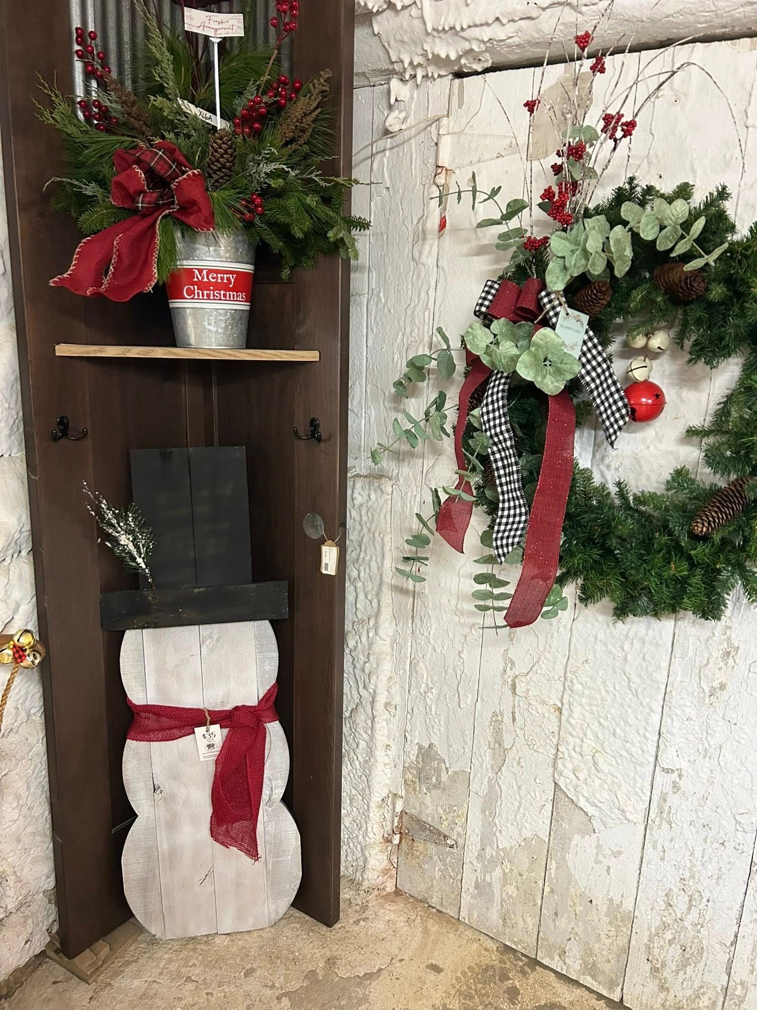 Rustic Christmas decorations in a corner: snowman, wreath, bucket of greenery with red bows.