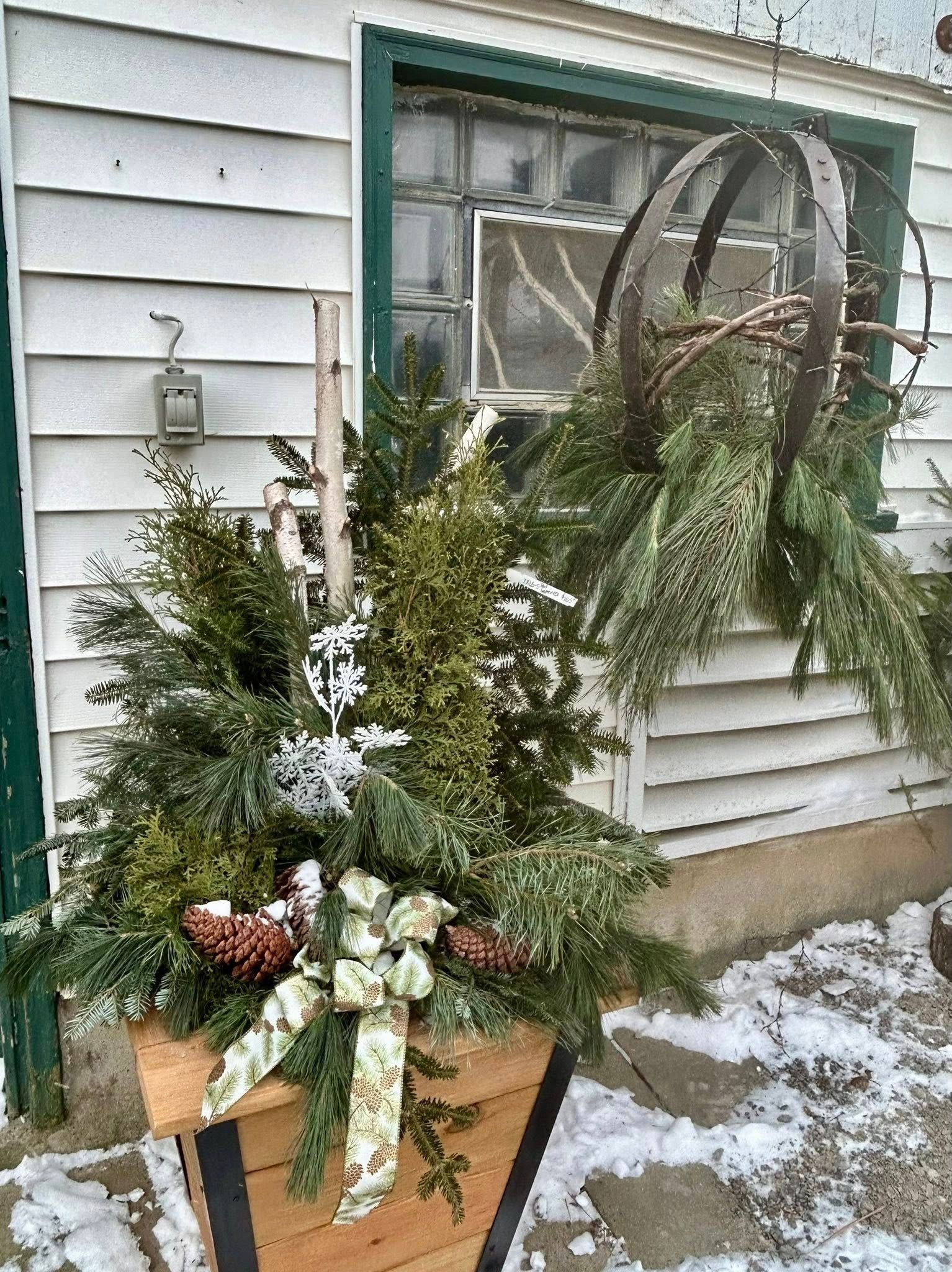 Wooden planter overflowing with winter greenery, against a white building with snow.