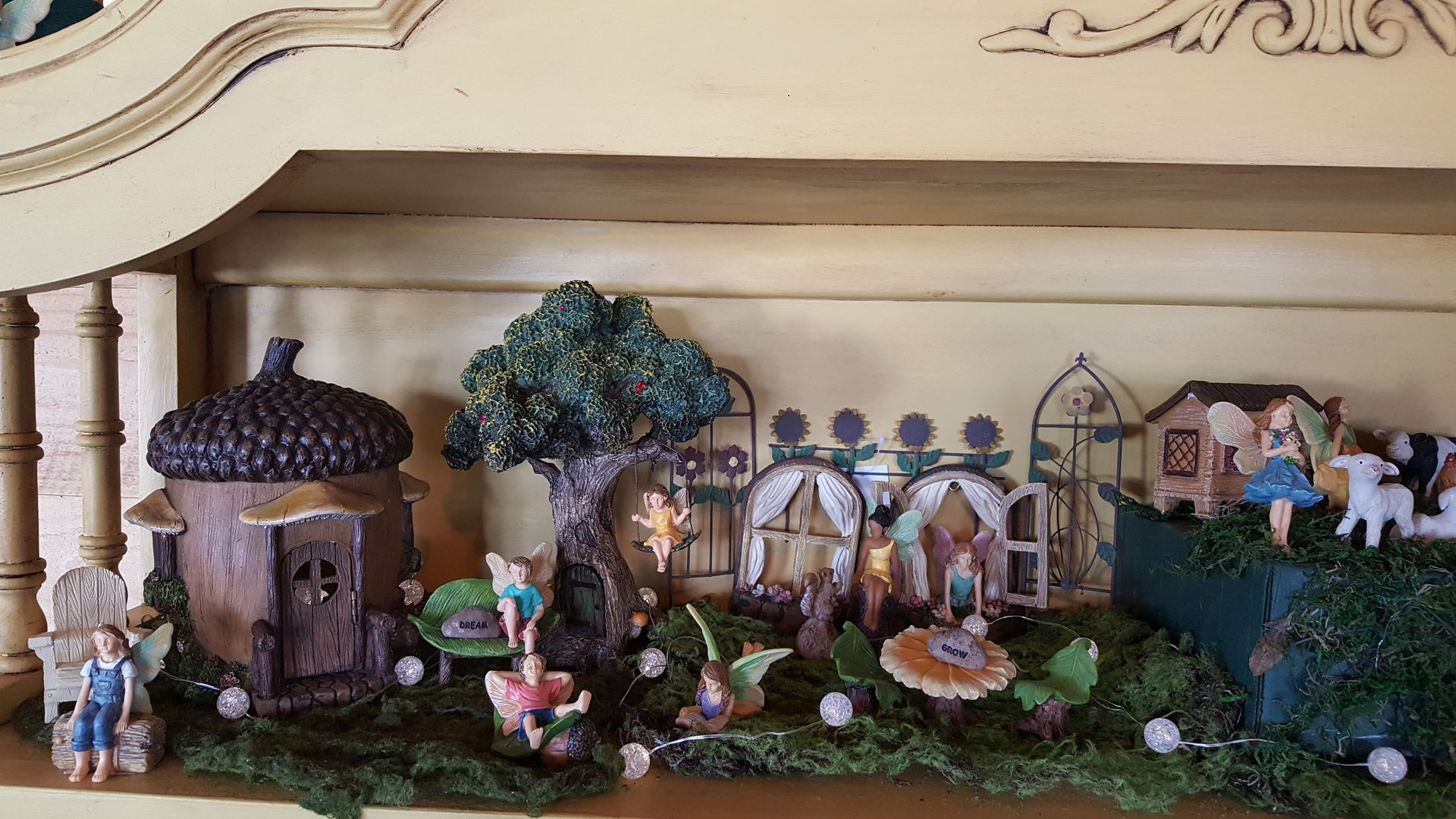 Miniature fairy garden display on a shelf, featuring whimsical houses, figures, and greenery.